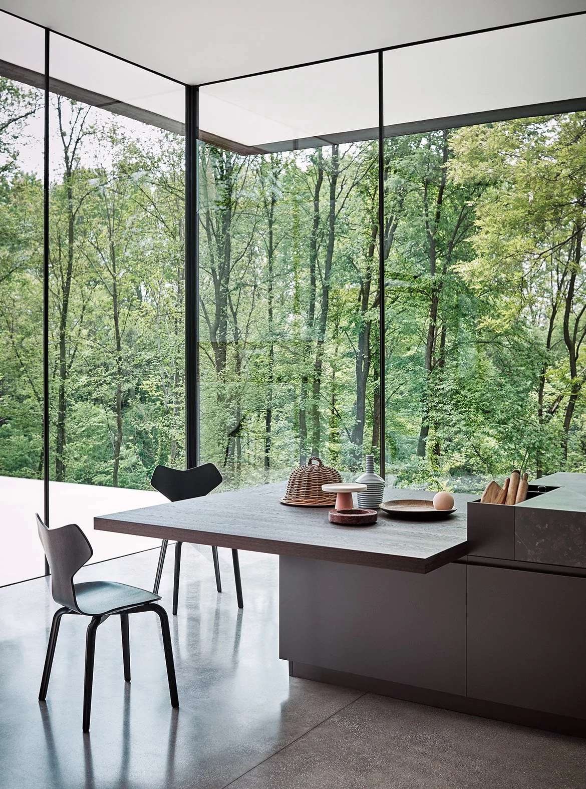 Innenansicht eines modernen Raumes mit großen Glaswänden, Blick auf einen grünen Wald, Esstisch mit Dekorationen, Stühle, steinartiger Küchenbereich.