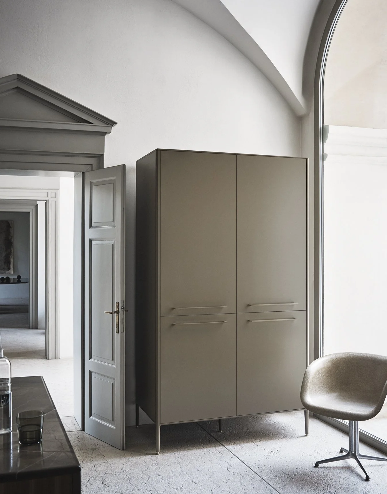 Moderner Raum mit grauem Kleiderschrank, einer weißen Tür, einem Stuhl und großen Fenstern, die viel Tageslicht hereinlassen.