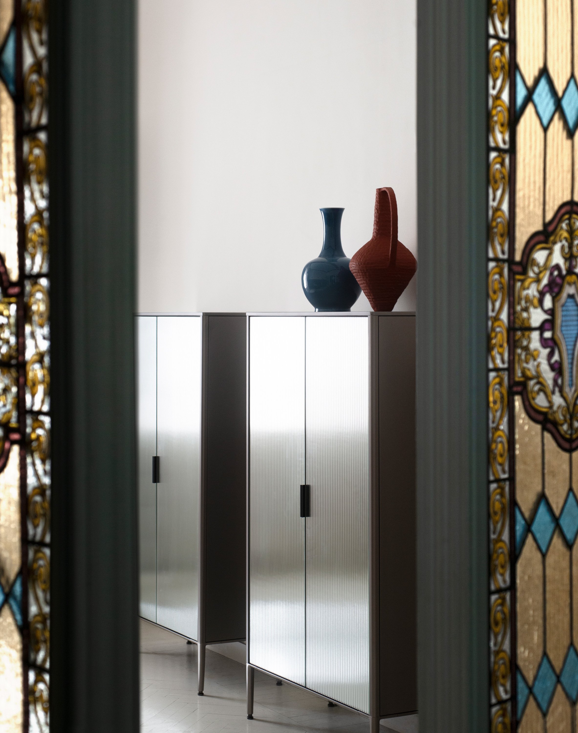 Zwei farbige Vasen, eine blaue und eine rote, stehen auf einem weißen Schrank vor einer weißen Wand. Das Bild ist durch einen offenen Türrahmen mit bunten, dekorativen Mustern eingerahmt.