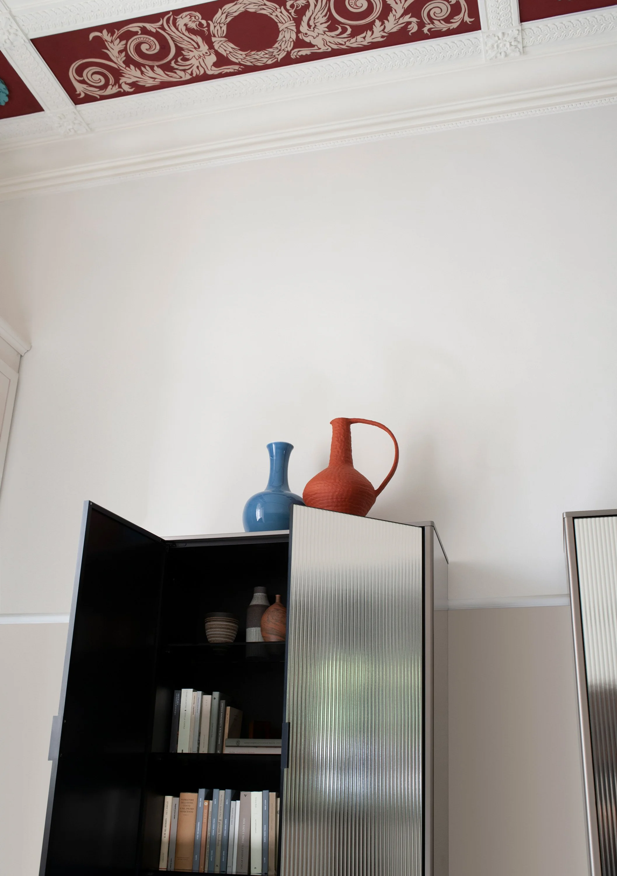 Ein Bücherregal mit schwarzen Schubladen und einer geschlitzten Glasschiebetür. Oben auf dem Regal stehen drei dekorative Vasen in Blau, Orange und Braun. Der Raum hat weiße Wände und eine Decke mit kunstvollen Schnitzereien.