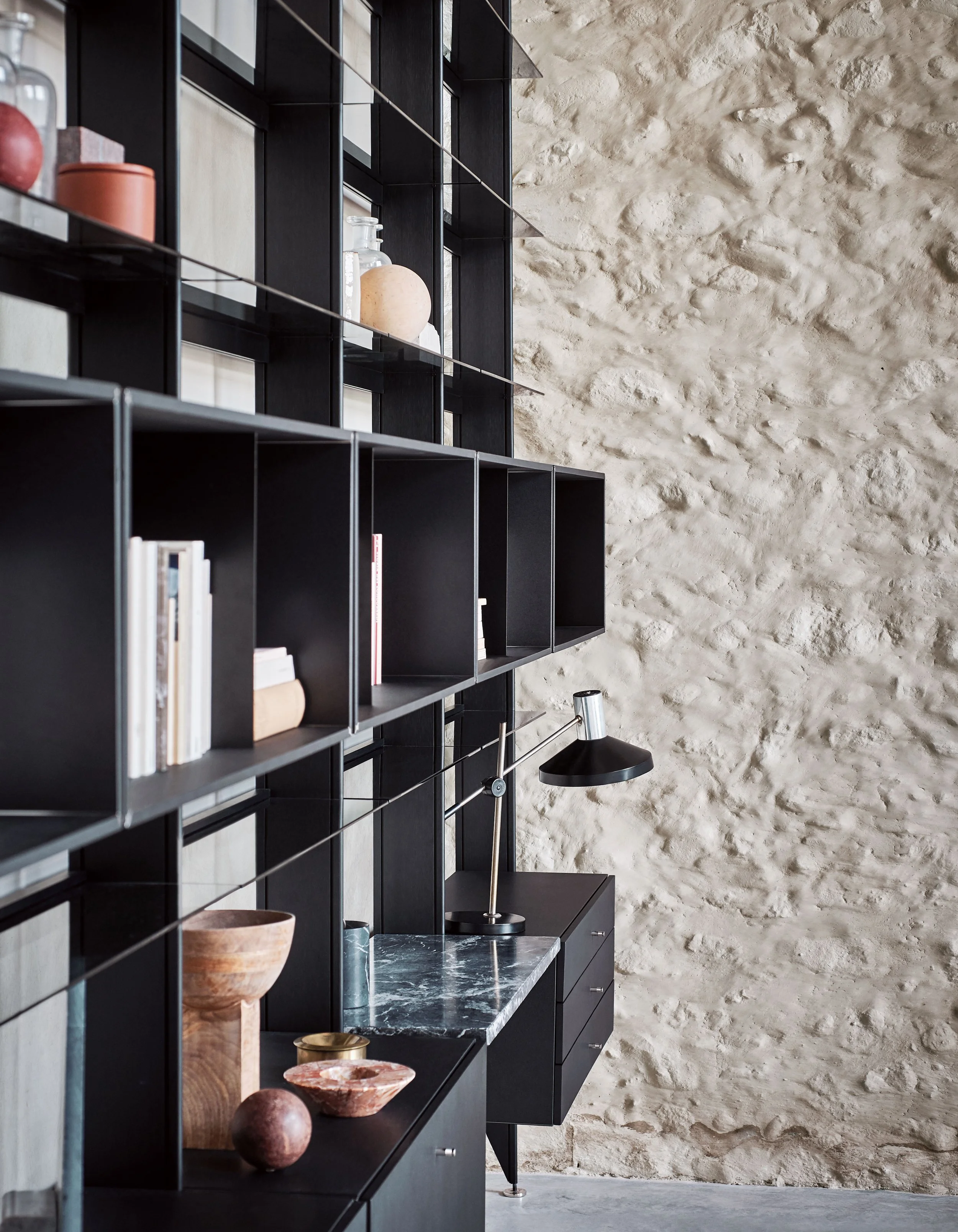 Schwarze Regale mit Dekorationsgegenständen, darunter Vasen, Bücher, Keramikschalen und Skulpturen, vor einer grob verputzten, hellen Steinwand in einem modernen Raum.