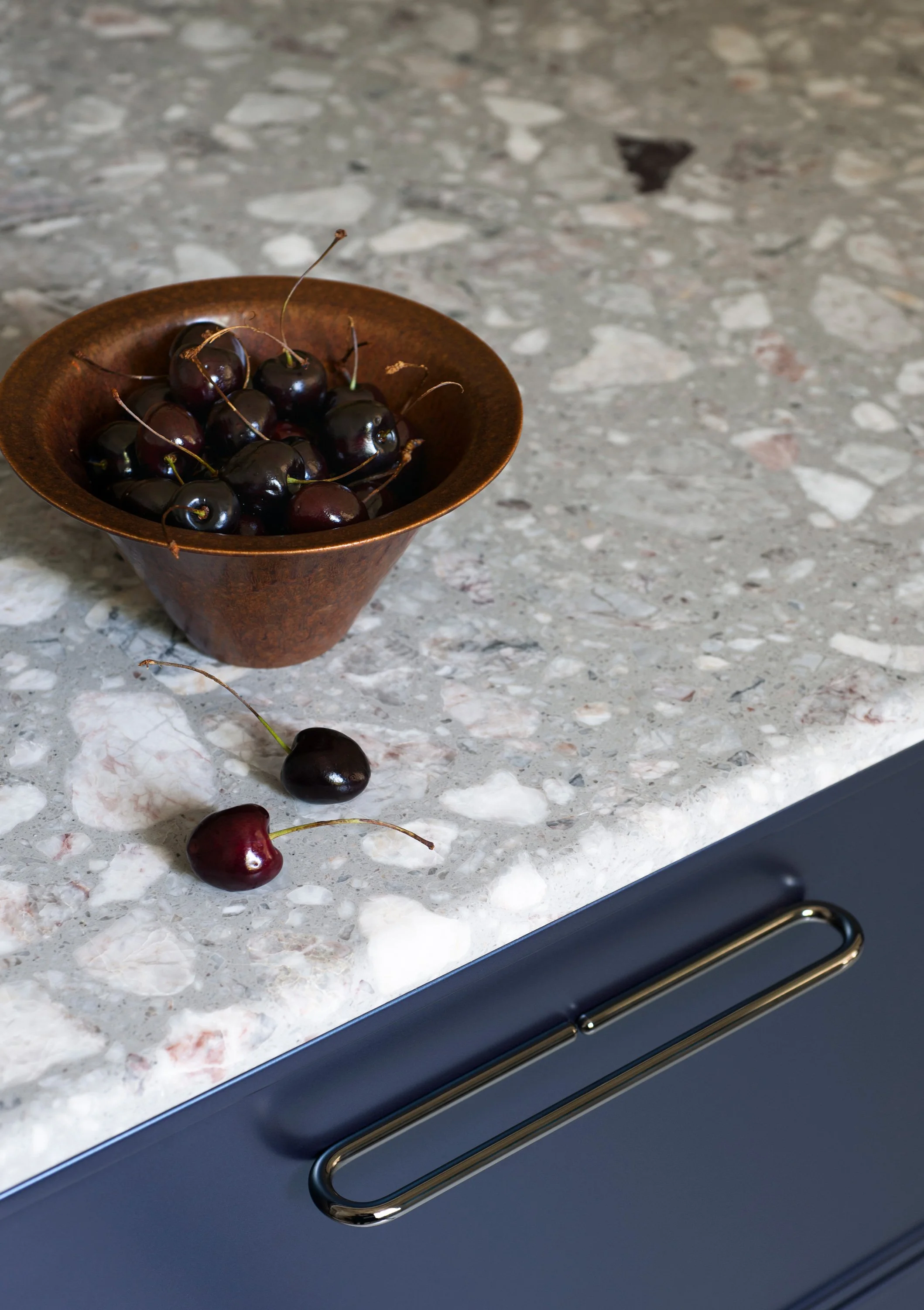 Eine Schüssel mit dunklen, reifen Kirschen auf einer grauen Marmortheke, einige Kirschen liegen daneben auf der Theke. Im Hintergrund sieht man eine dunkle Küchenschublade.