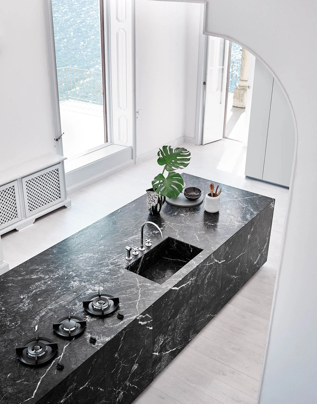 Moderne Küche mit einer schwarzen Marmorarbeitsplatte, einem Wasserhahn, einem kleinen Waschsieb, einer Pflanze, Schalen und Küchenutensilien. Offene Türen leading zu einem Balkon mit Blick auf Wasser.