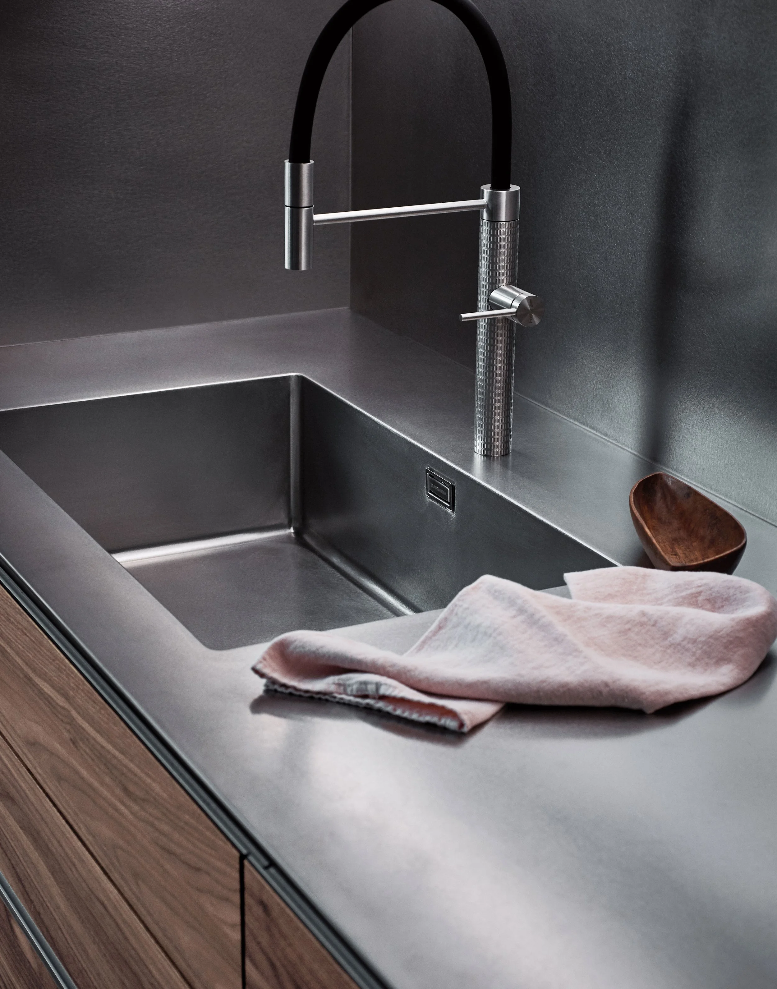 Moderner Edelstahlspüllbecken mit schwarzem Wasserhahn, Krümel und ein Waschlappen liegen auf der Ablage, eine Holzschale steht neben dem Waschbecken.