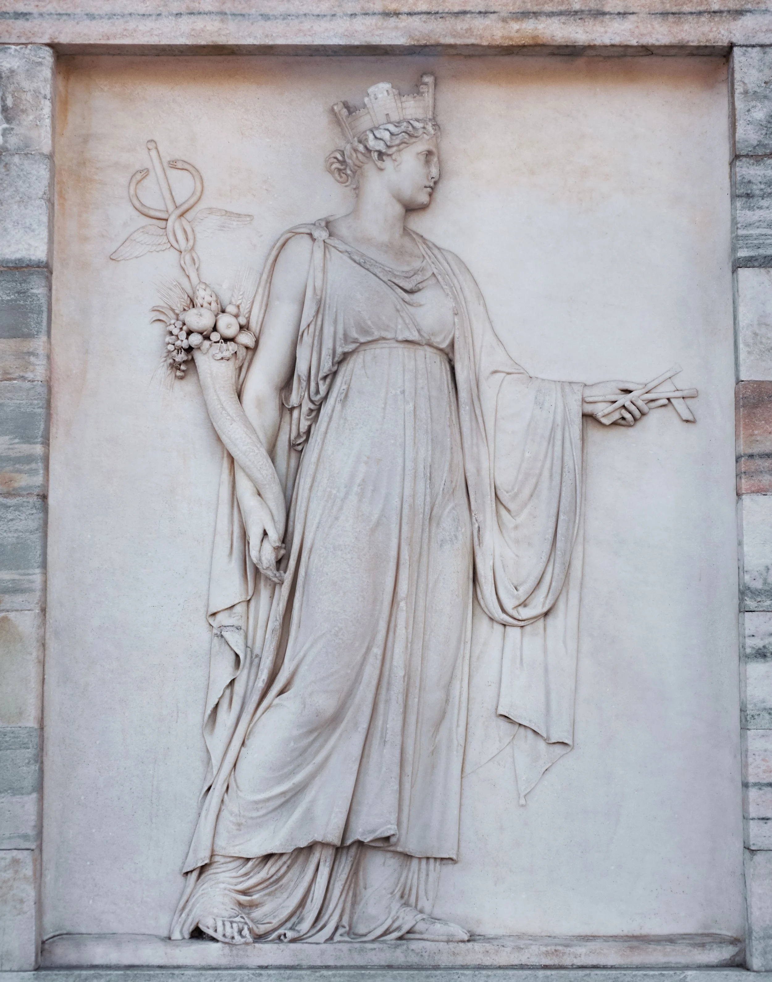 Relief eines Kopfes einer Frau mit Krone, die einen Stab und ein Schriftstück hält.