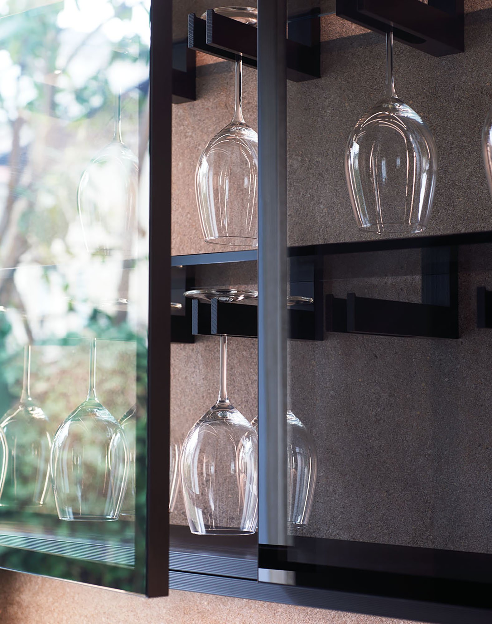 Transparent Weingläser hängen kopfüber an einer Metallhalterung in einem Regal, mit einer Fenster und grünen Bäumen im Hintergrund.