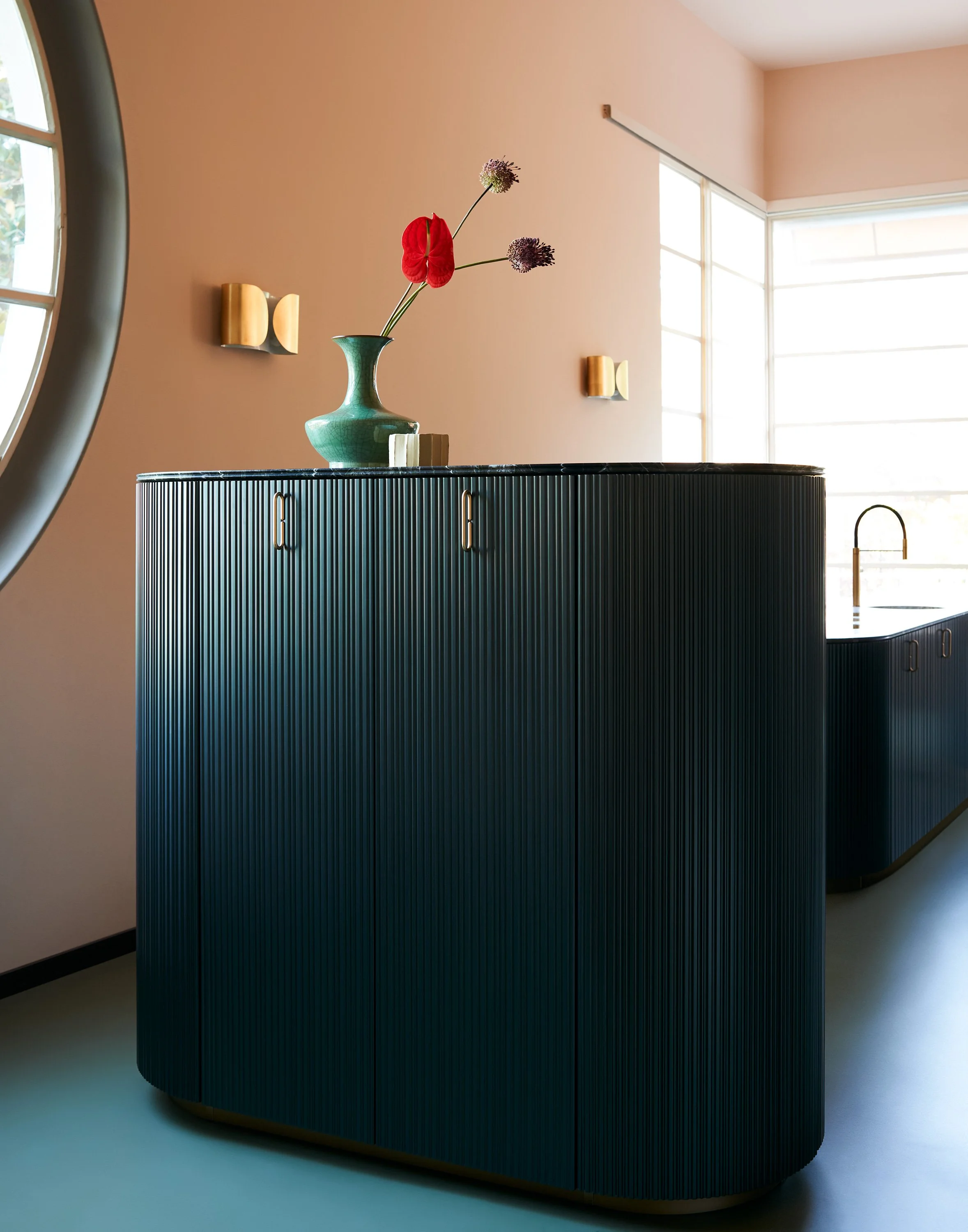 Moderner, dunkelblauer Schrank mit vertikalen Linien und goldenen Griffen, auf dem eine grüne Vase mit bunten Blumen steht, im Hintergrund eine große Fensterfläche und beige Wände.