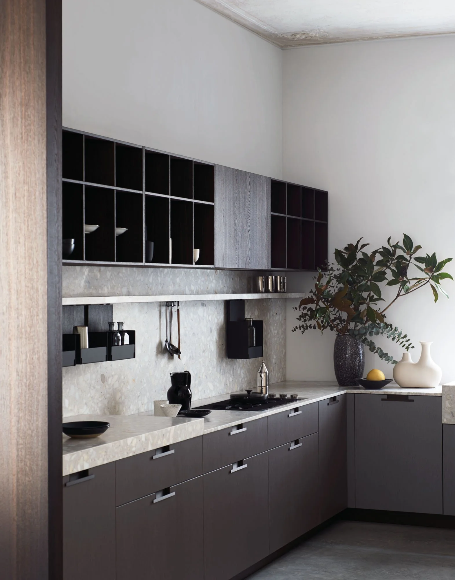 Moderne Küche mit grauen Schränken, weißer Steinarbeitsplatte, schwarzen Geschirrhaltern, einer großen Vase mit grünen Zweigen, schwarzer Kaffeemaschine, dunklen Geschirr, Blumentopf, hellgrauer Wand und dunklem Boden.