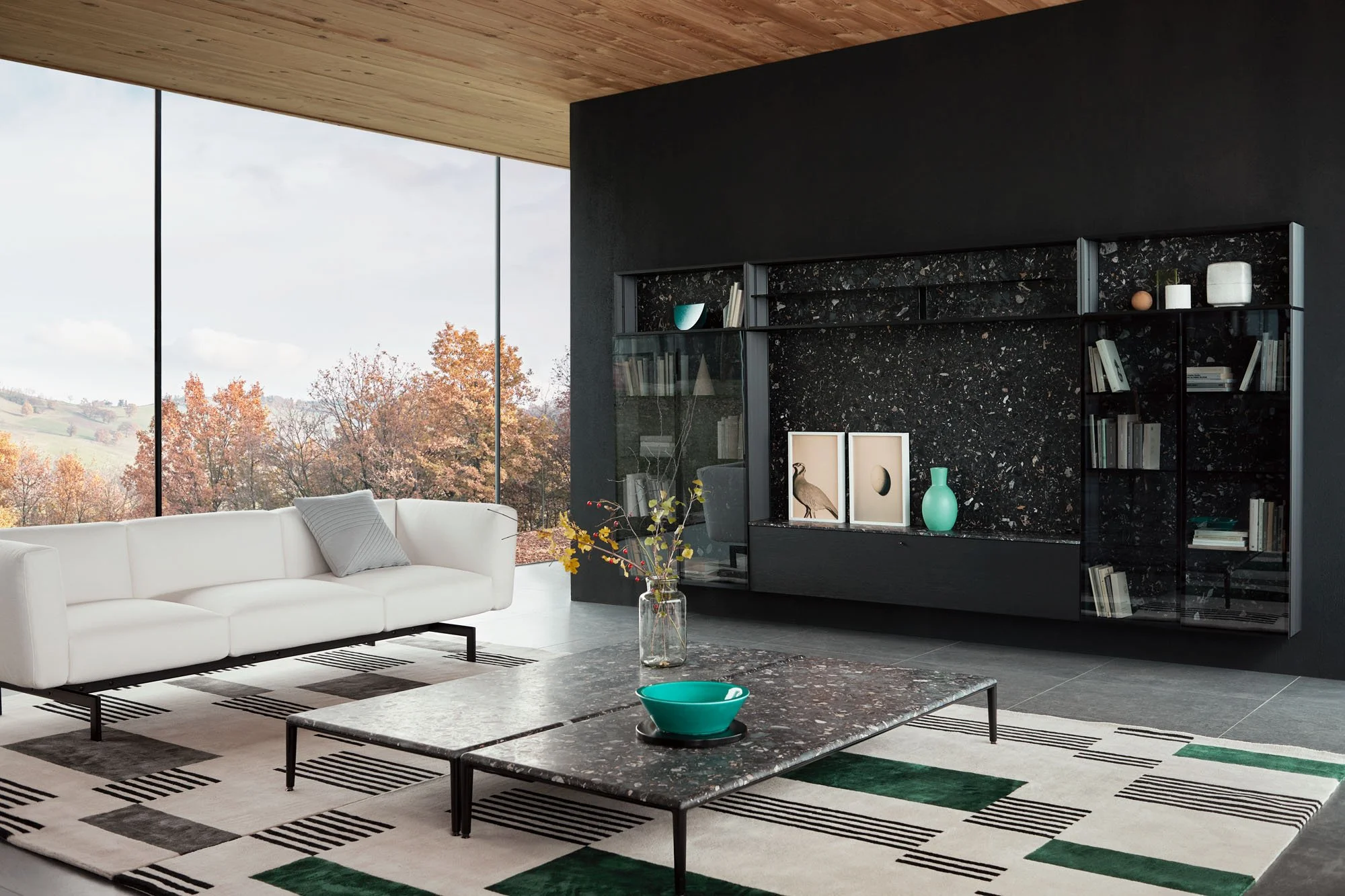 Modernes Wohnzimmer mit großer Fensterfront, einer weißen Couch mit grauem Kissen, einem schwarzen Regal mit Dekorationen und Büchern, einer schwarzen Marmortisch, einem Glasvase mit Blumen, einer grünen Schale und einem Teppich mit schwarz-weißen Mu