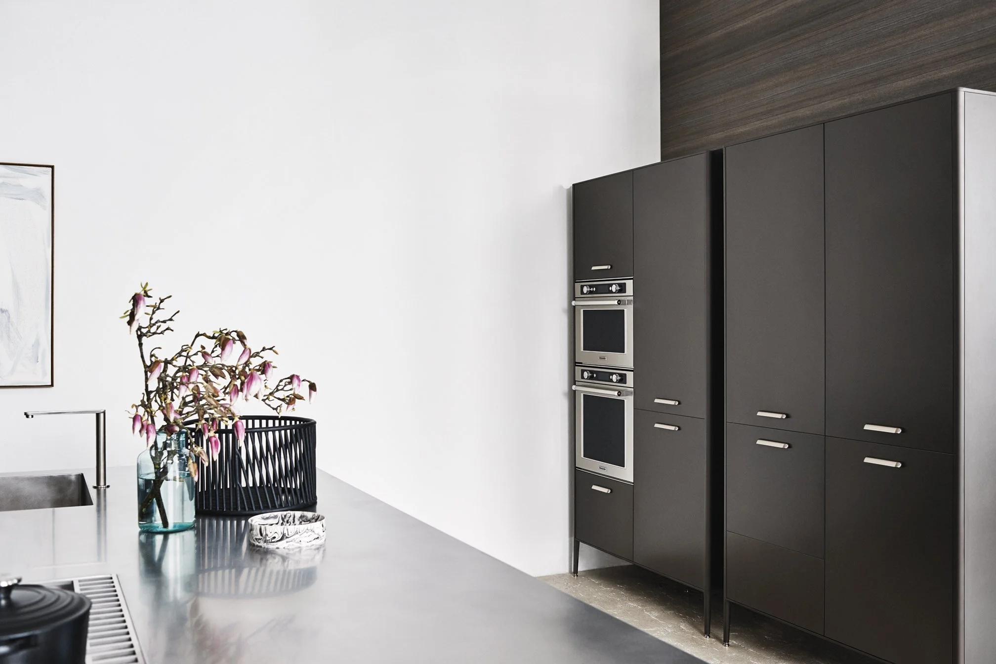 Moderner Küchenraum mit schwarzen Schränken, Backofen, Kochfeld und Geschirrspüler. Es gibt einen weißen Tisch mit einer blauen Vase mit Blumen. Eine Wand ist weiß, die andere hat eine Holzverkleidung.