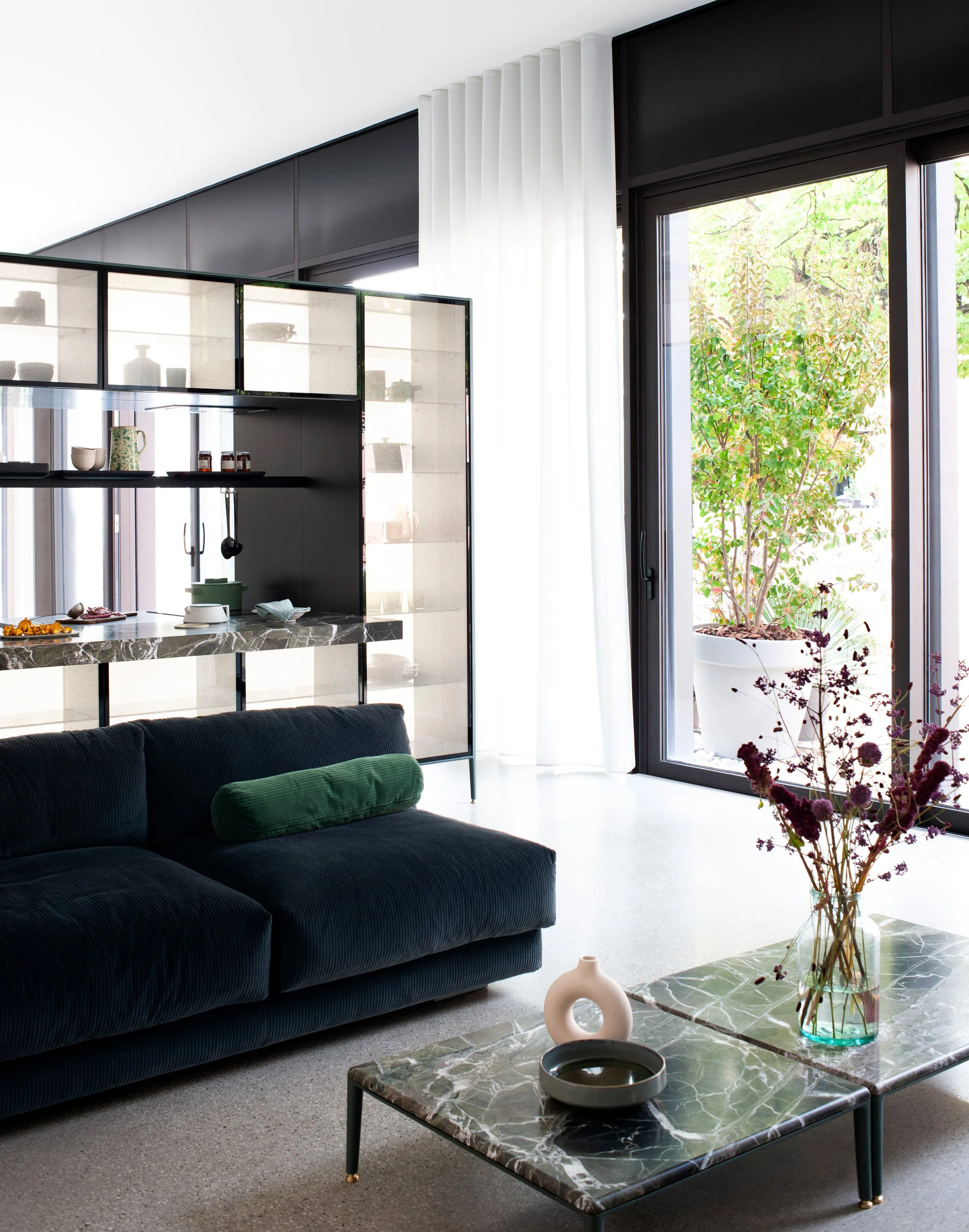 Ein modern eingerichteter Essbereich mit schwarzem Sofa, grünem Polster, Marmor-Couchtisch mit grüner Maserung, dekorativer Vase mit Blumen und großem Fenster mit Blick auf einen Garten mit Bäumen.