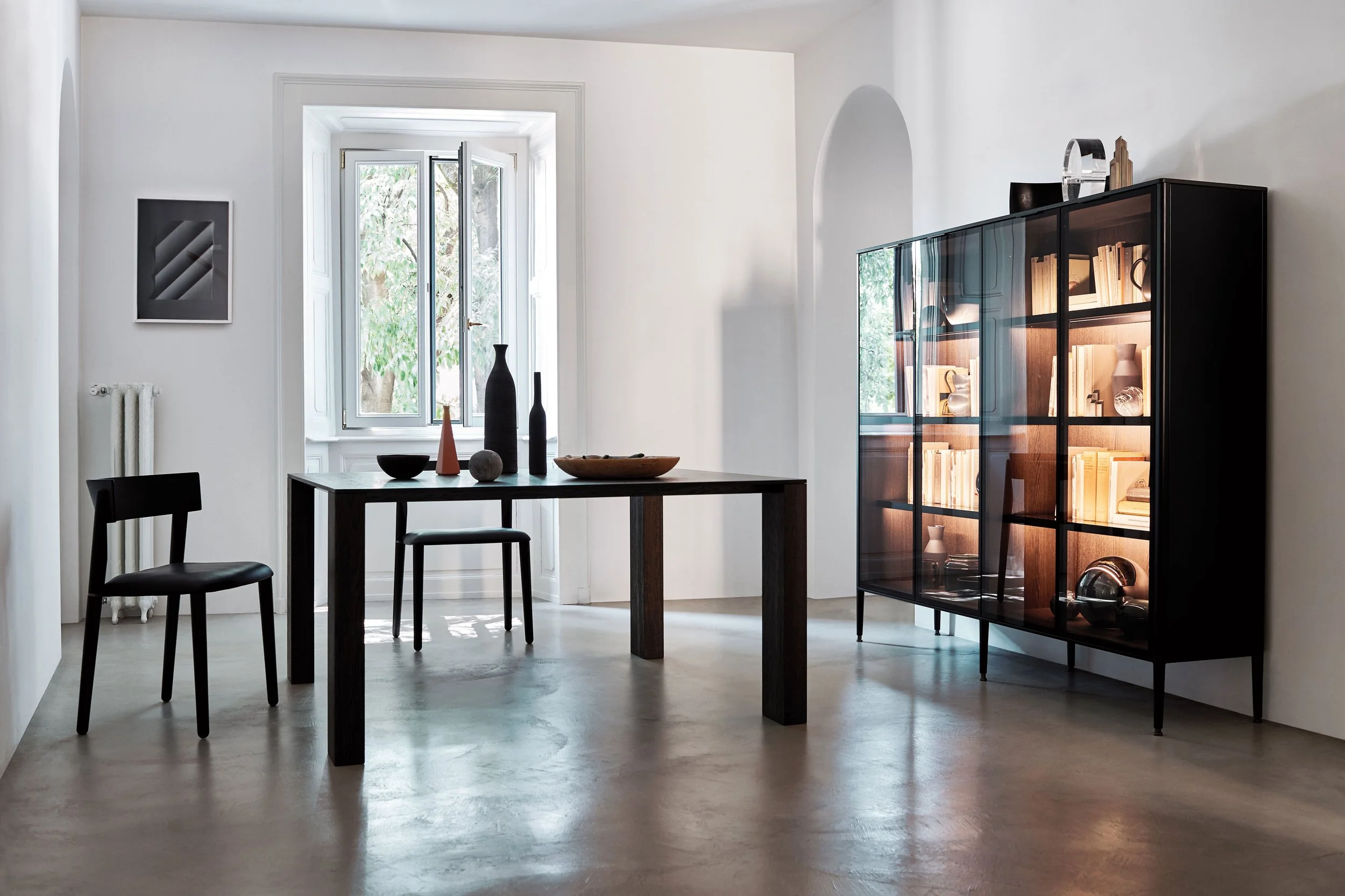 Ein modern eingerichtetes Wohnzimmer mit einem großen Fenster, einem Esstisch aus Holz, schwarzen Stühlen und einem schwarzen Vitrinenschrank mit Glastüren, der Bücher und dekorative Gegenstände enthält.