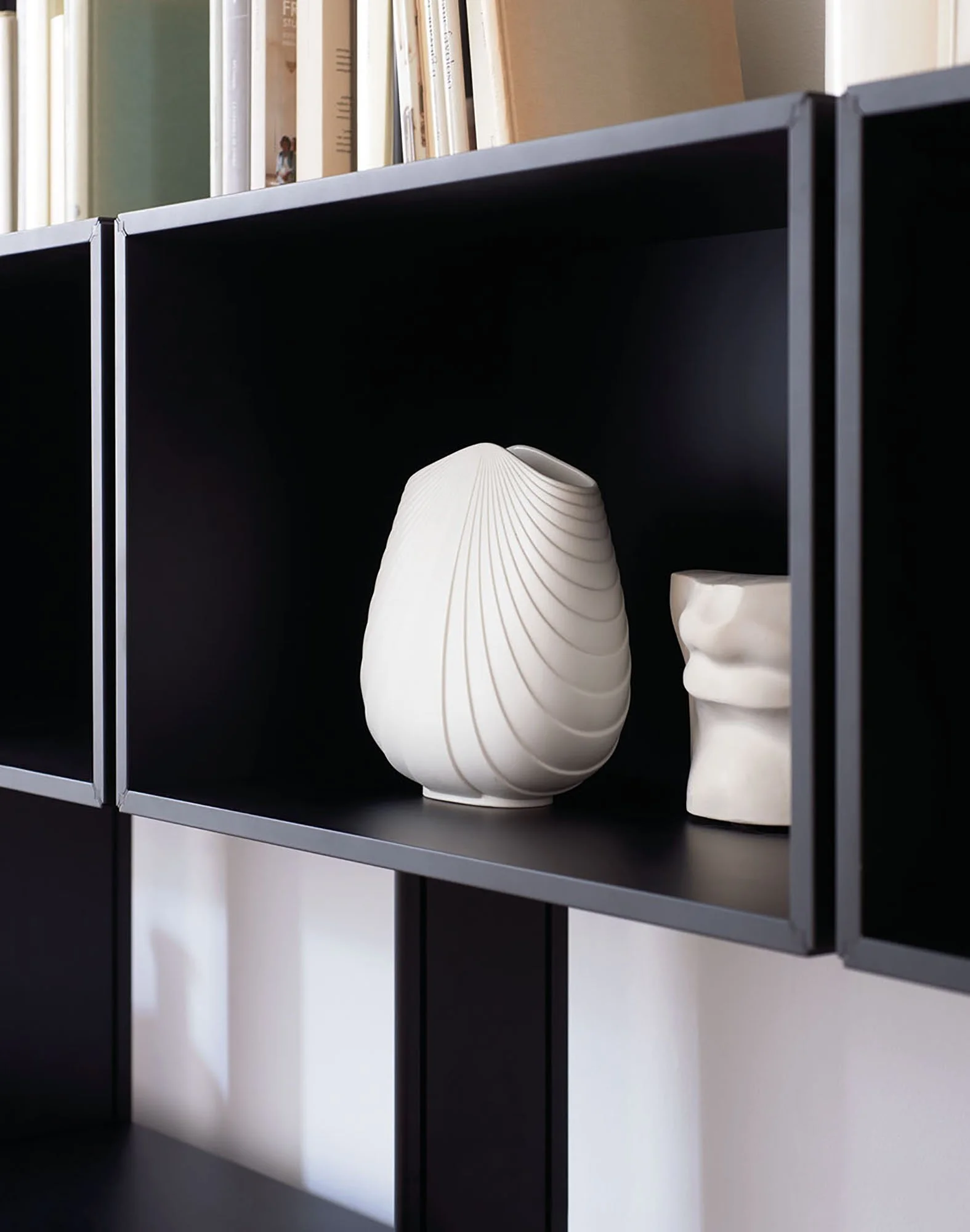 Schwarzes Regal mit weißen Keramikvasen und einer Bücherwand im Hintergrund.
