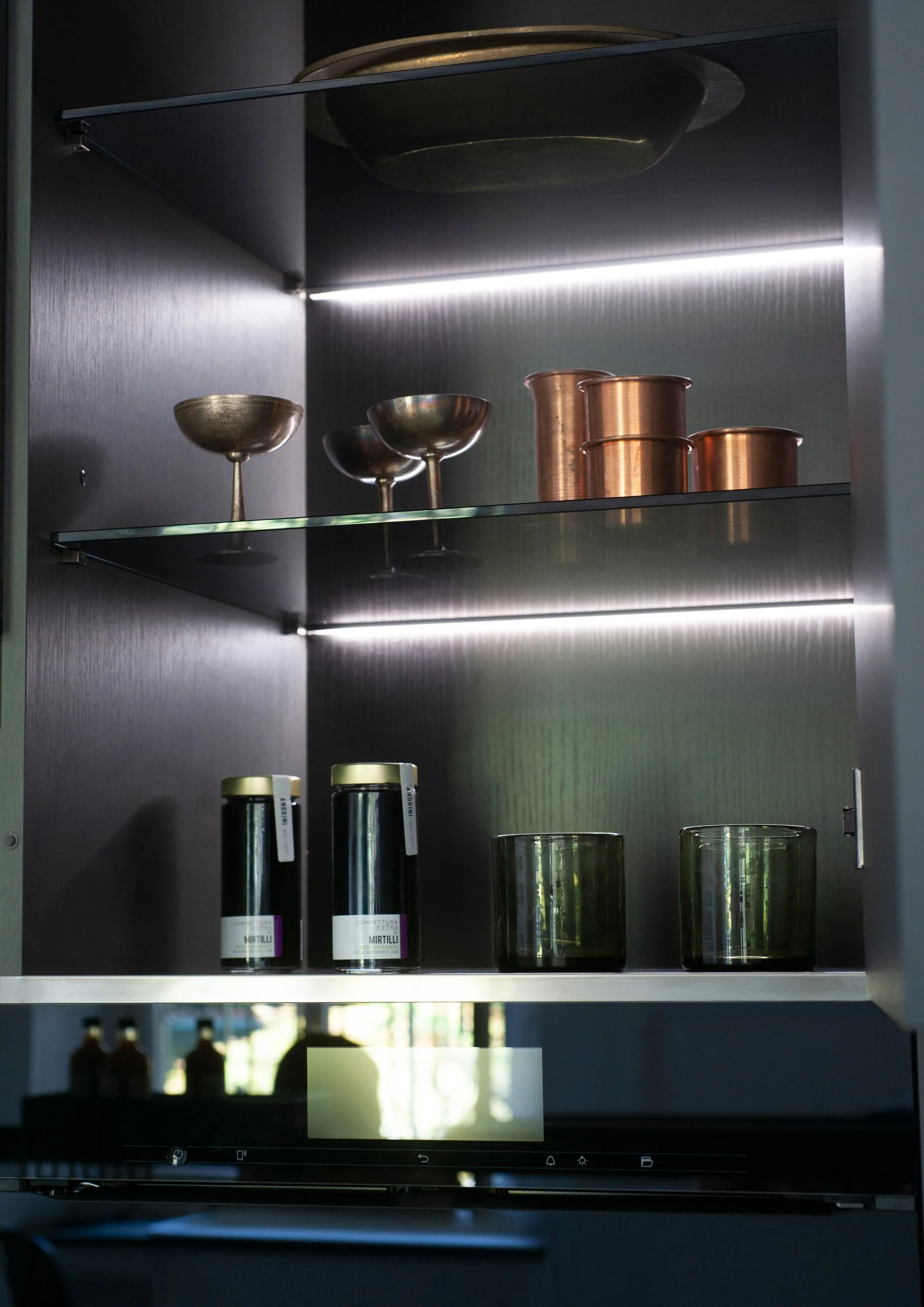 Blick in einen Küchen-Schrank mit drei Regalen, die stilvolle Küchenutensilien und Gewürzgläser enthalten, mit Blick auf eine helle Küche im Hintergrund.