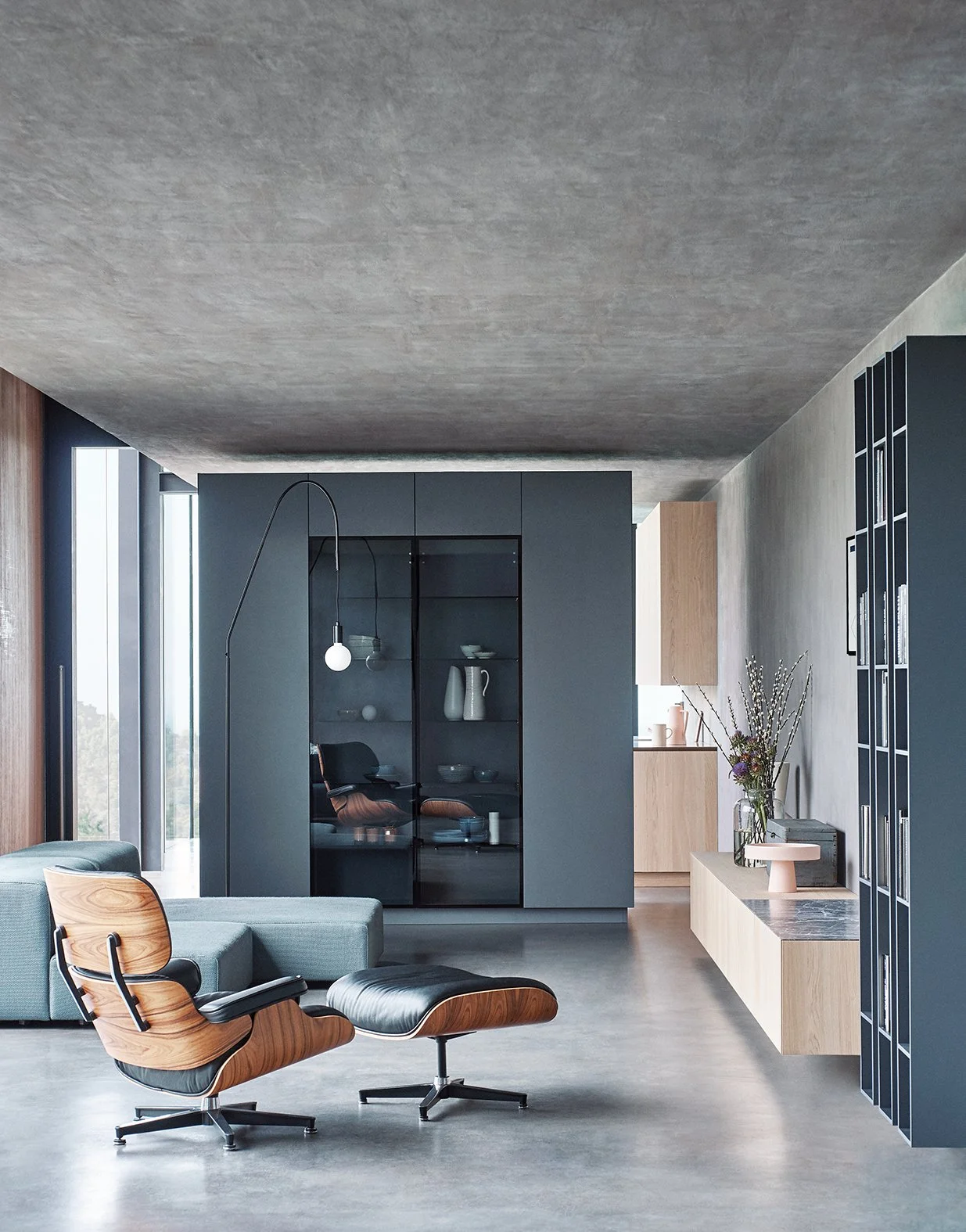 Moderner offene Wohnraum mit grauer Couch, Holzstuhl, und schwarzen Vitrinen in einem hellen, minimalistischen Raum, mit Blick aus großen Fenster