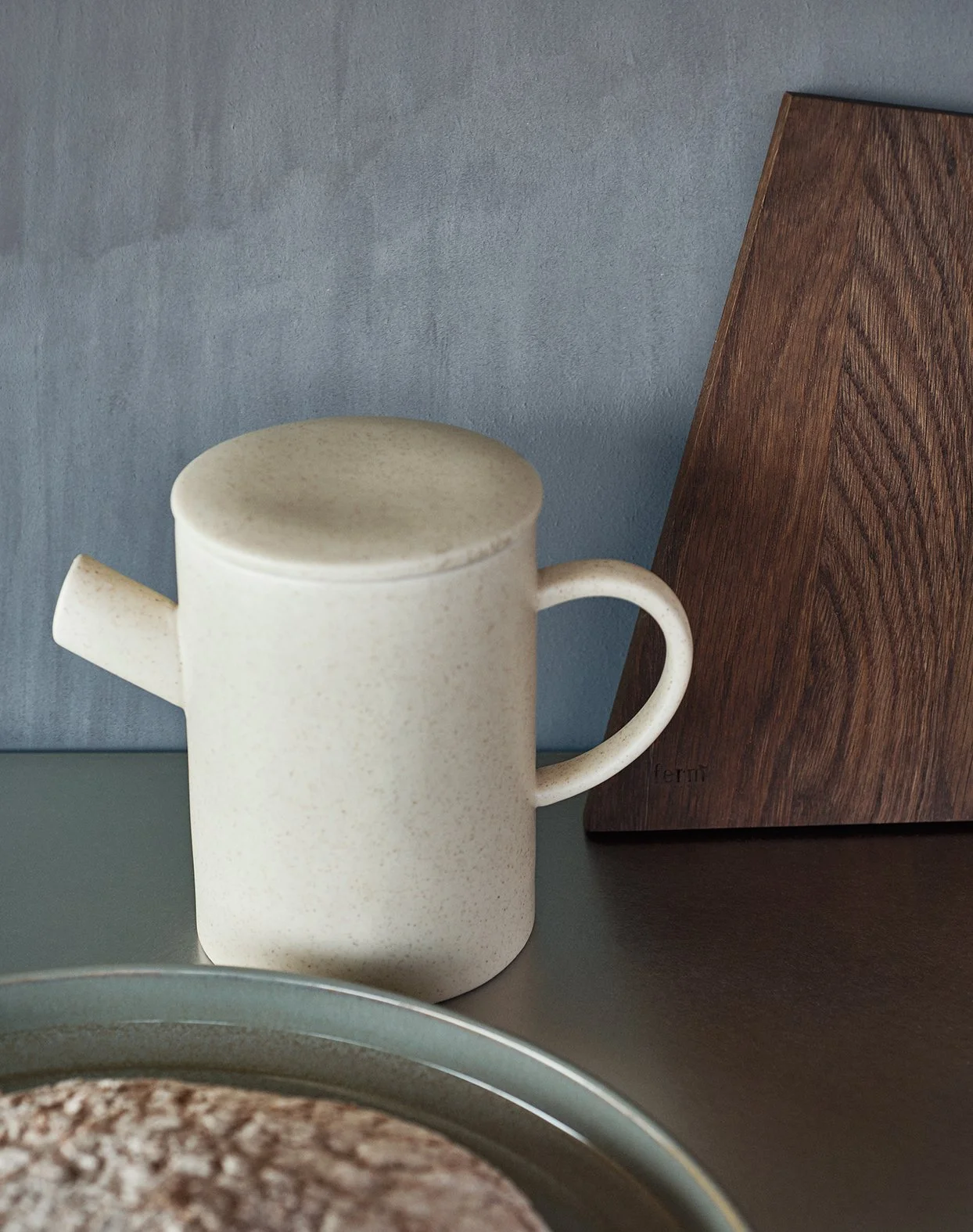 Ein beigefarbener Kaffeekannen-Topf steht auf einem Tisch, im Hintergrund ein hölzernes Schneidebrett und eine graue Wand.