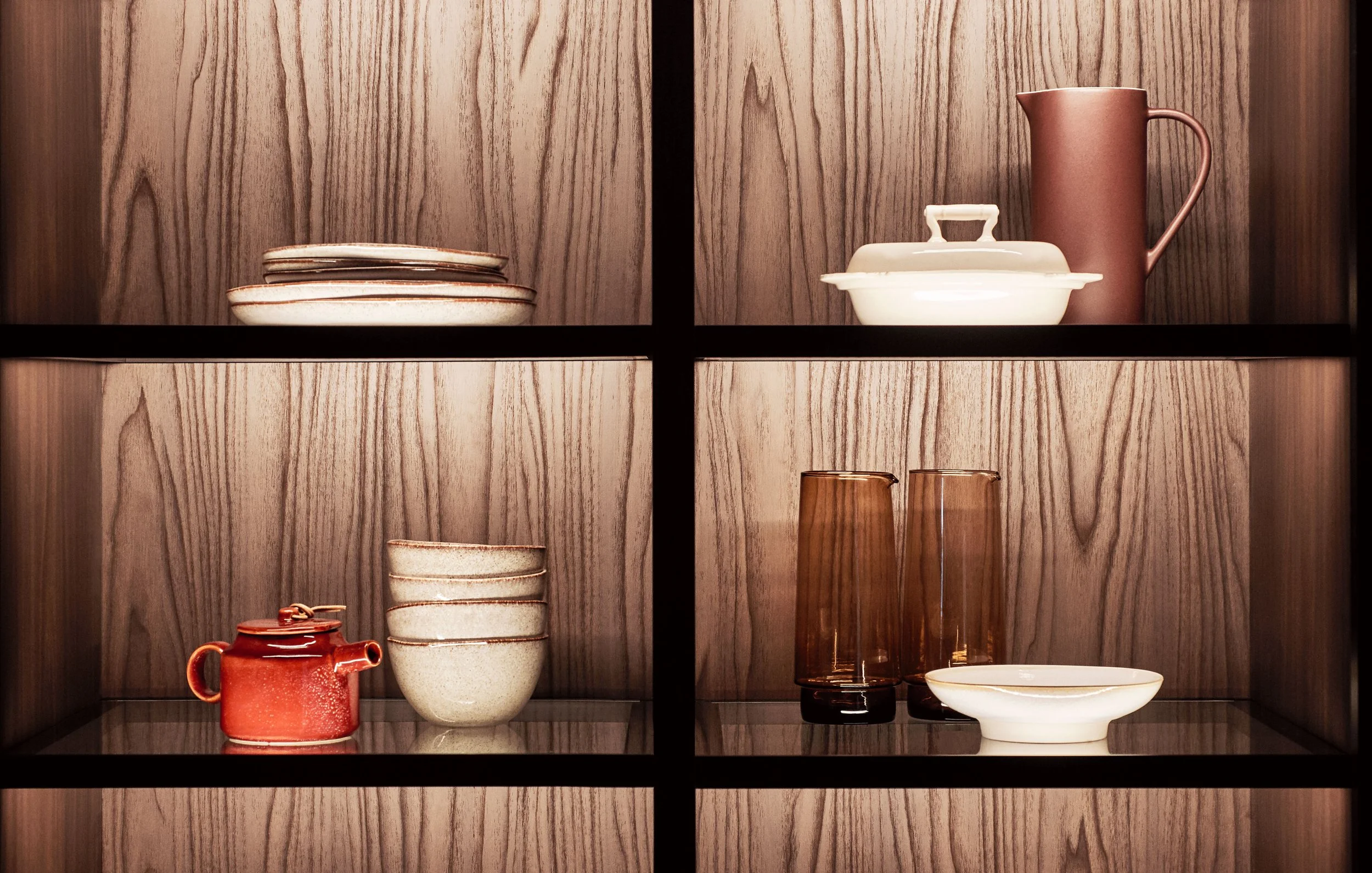 Auf einer Holzwand befinden sich Keramik- und Glaswaren in einem Schrank mit schwarzen Rahmen, darunter eine rote Teekanne, weiße und braune Tassen, eine weiße Schüssel, eine braune Glaskaraffe und eine weiße Schale.