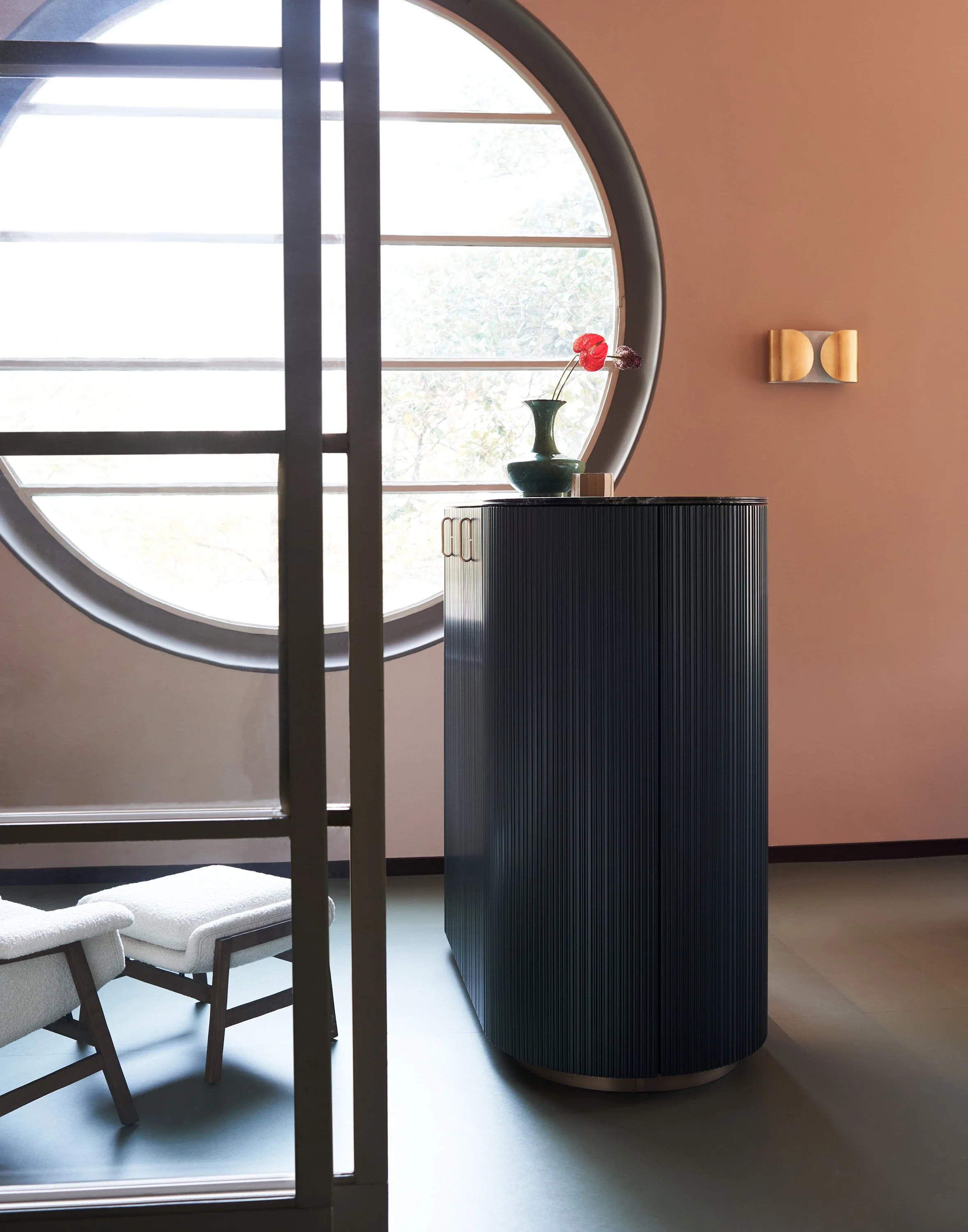 Ein helles Wohnzimmer mit einem großen runden Fenster, einem schwarzen Möbelstück mit einer Vase und einer Blume sowie einem weißen Stuhl.