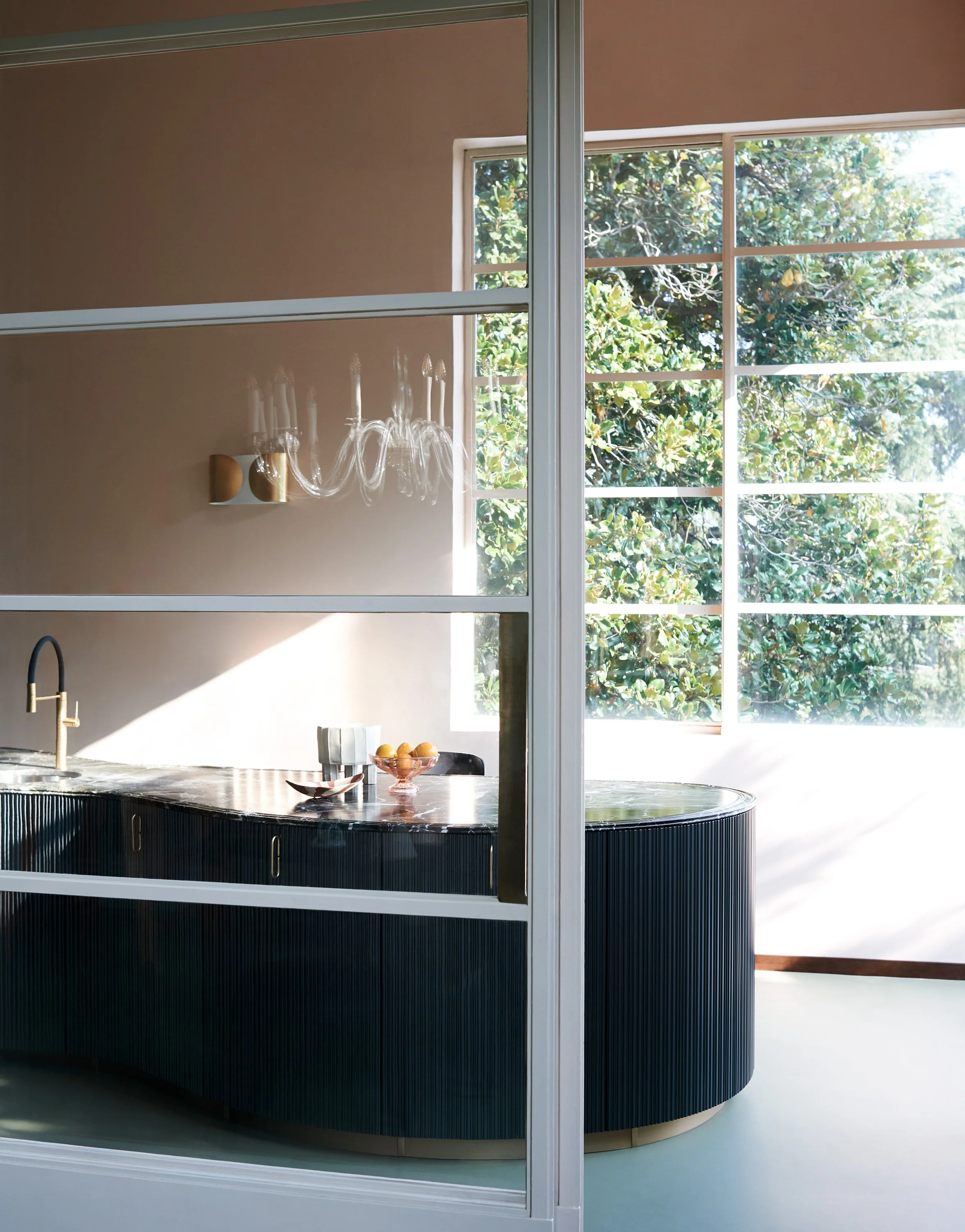 Innenraum mit schwarzer Kücheninsel, ein Radiator, eine Schüssel mit Orangen und ein Wasserkocher, große Fenster mit Blick auf grüne Bäume, helle Atmosphäre.