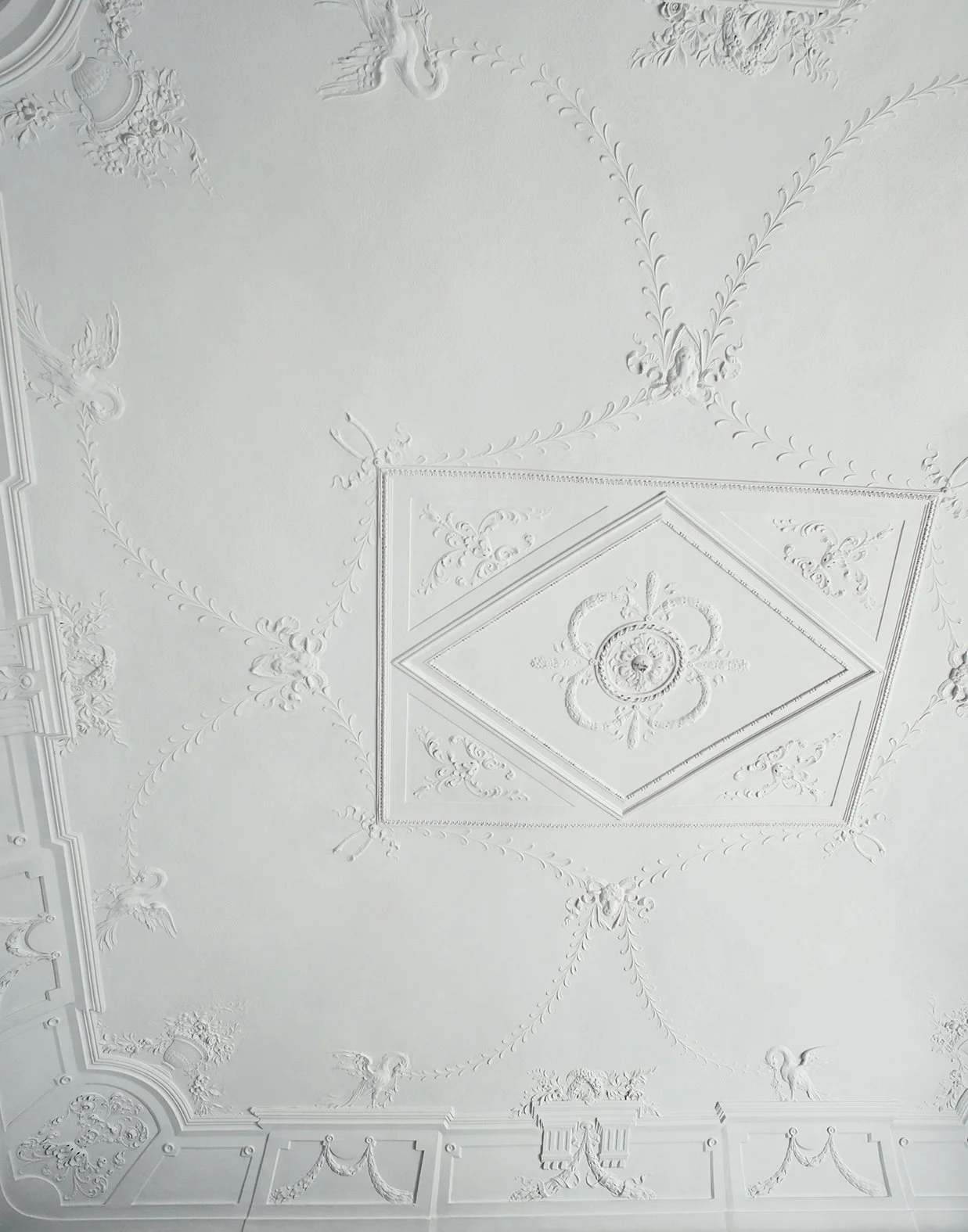 Weiße Decke mit aufwendigem, kunstvollem Stuckmuster, das zentrale geometrische Muster, Flügel, Blumen und andere ornamentale Details zeigt.