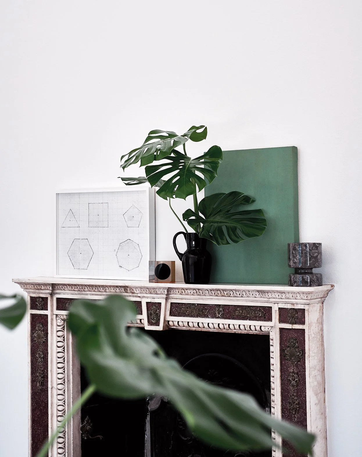 Ein antiker Kamin mit dekorativen Details, darauf eine schwarze Vase mit großen grünen Monstera-Blättern, umgeben von Kunstwerken und Dekorationsgegenständen in minimalistischer Gestaltung.