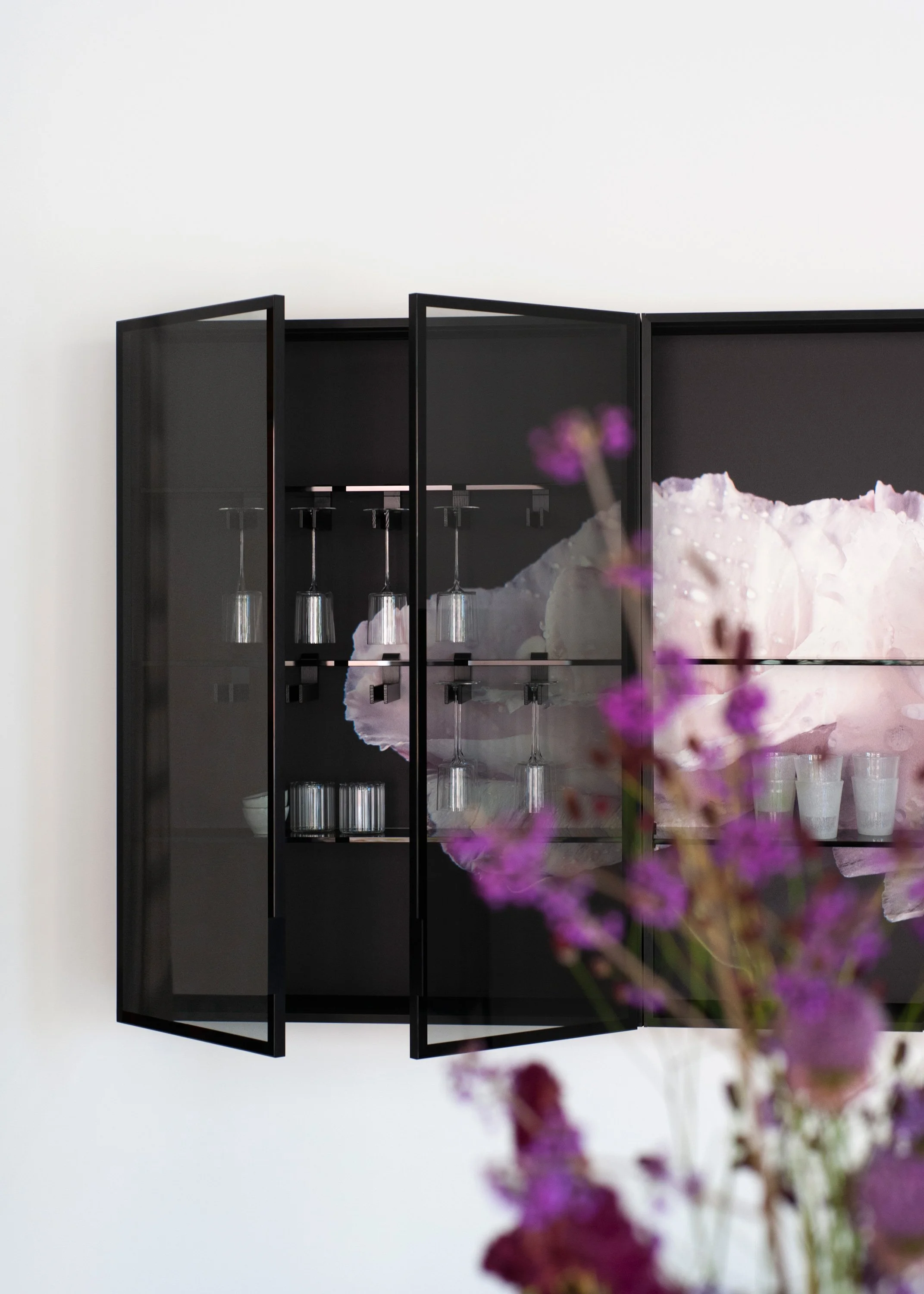 Ein schwarzer Geschirrspüler mit geöffneten Türen, darin sind Gläser und Geschirr, vor einem weißen Hintergrund, im Vordergrund unscharf Blumen in Violett und Pink.