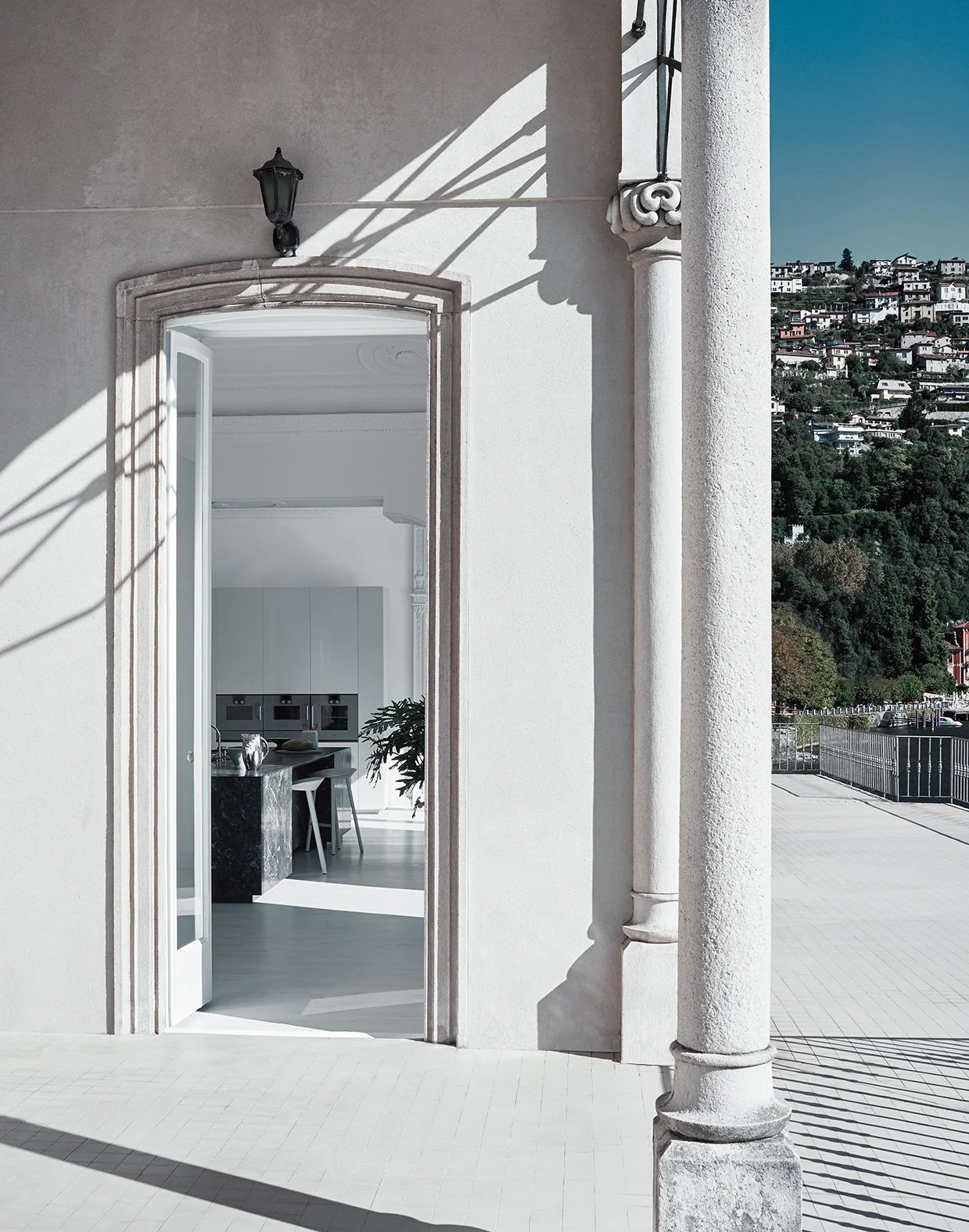 Ein Blick durch eine offene Tür in ein modernes, helles Innere mit weißer Küche und einem schwarzen Küchenblock. Draußen ist eine Terrasse mit Weitblick auf eine hügelige Wohngegend. Die Hausfassade ist aus hellem Putz mit einer klassischen Säule und