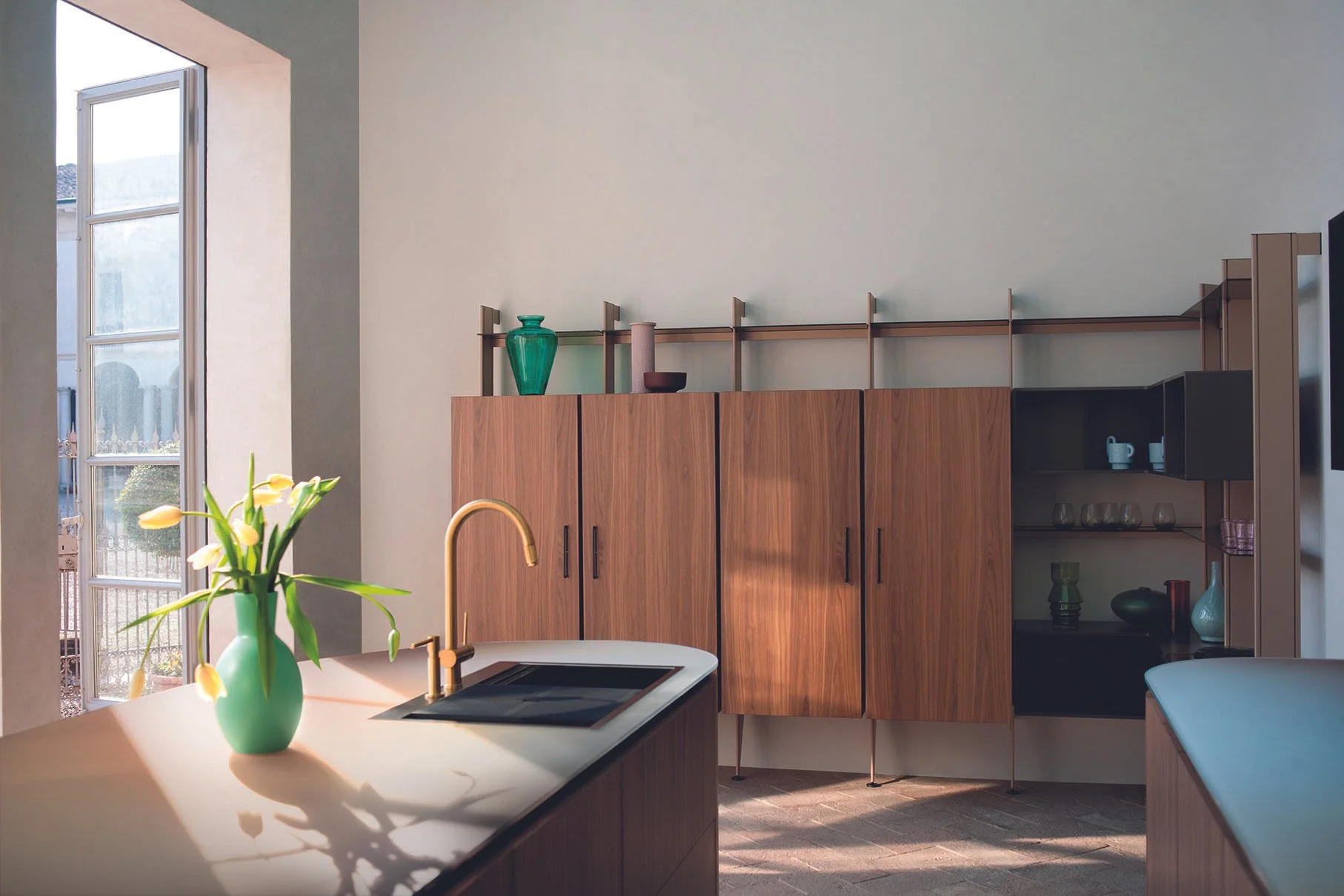 Eine moderne Küche mit Holzschränken, einer weißen Arbeitsfläche, einem Wasserhahn und einer Vase mit gelben Blumen auf der Theke. Fenster mit Blick nach draußen bringen natürliches Licht in den Raum.