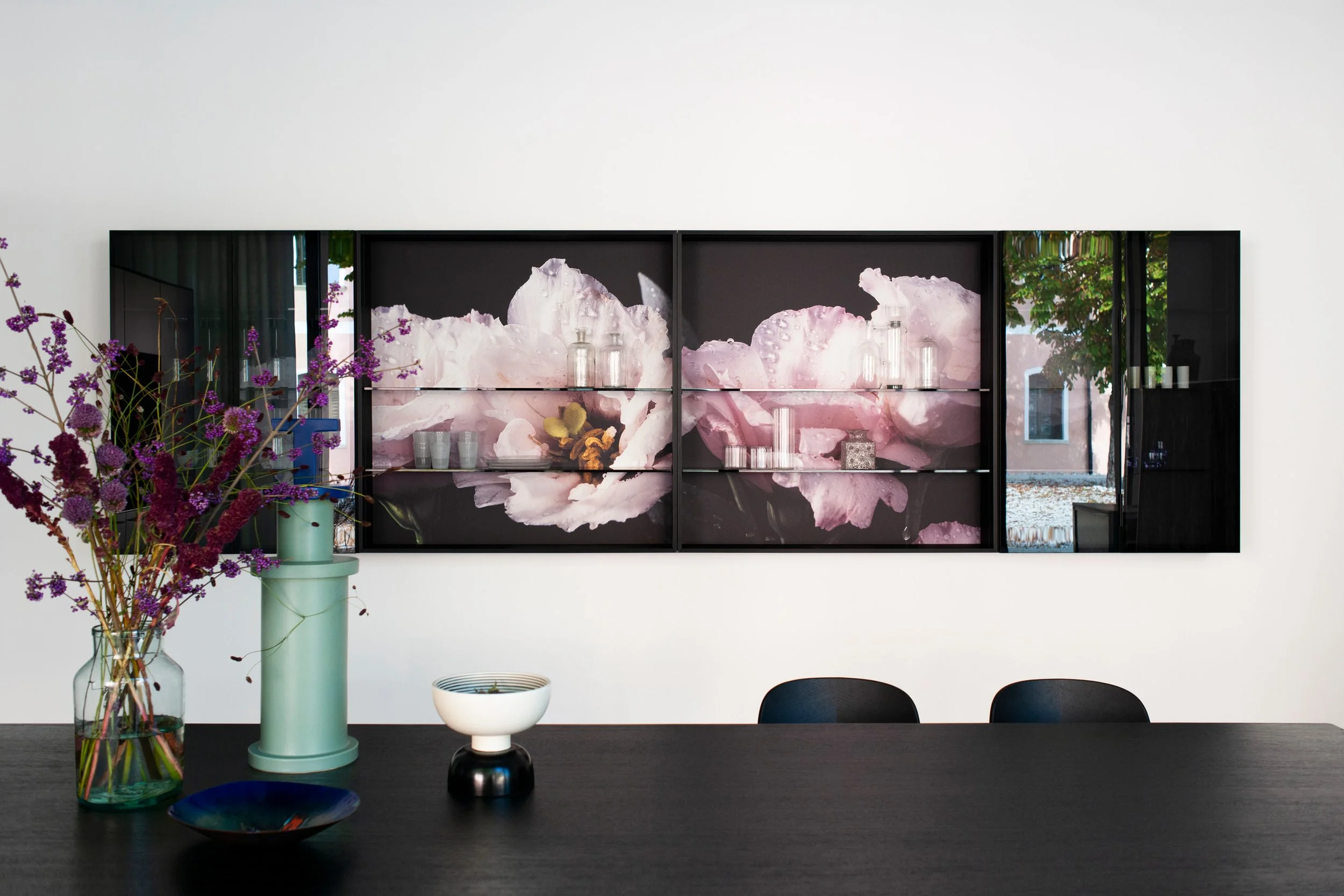 Ein Wohnzimmer mit einer schwarzen Kommode, auf der sich dekorative Gegenstände wie kleine Glasflaschen, eine Blume im Topf und eine Musterbox befinden. An der Wand hängt ein großformatiges Kunstwerk mit rosa und weißen Blumenmotiven, umgeben von ein