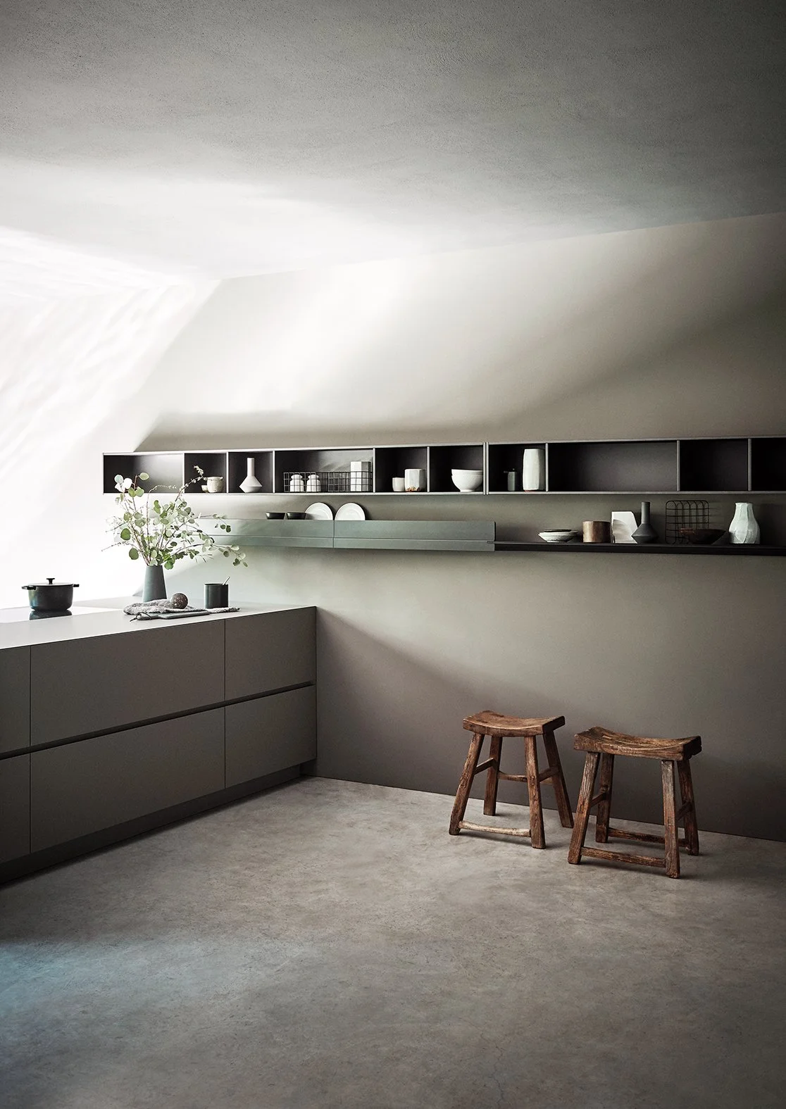 Moderner, minimalistischer Küchenraum mit grauen Schränken, offenen Regalen mit Geschirr und Dekoration, zwei alte Holzschemel, eine Vase mit grünen Zweigen auf der Arbeitsplatte und weiche Beleuchtung.