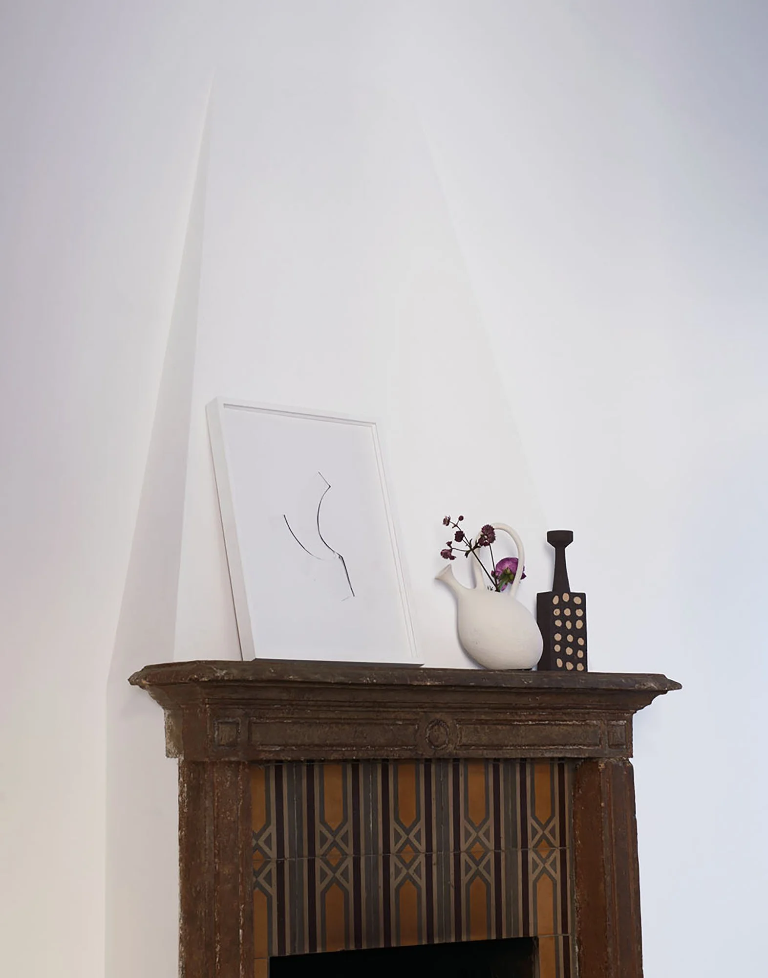 Ein antiker Kamin mit Holzblock, darüber eine Wanddeko mit weißen Vasen, einer dunklen Vase und einer Vase mit roten Blumen, auf weißer Wand