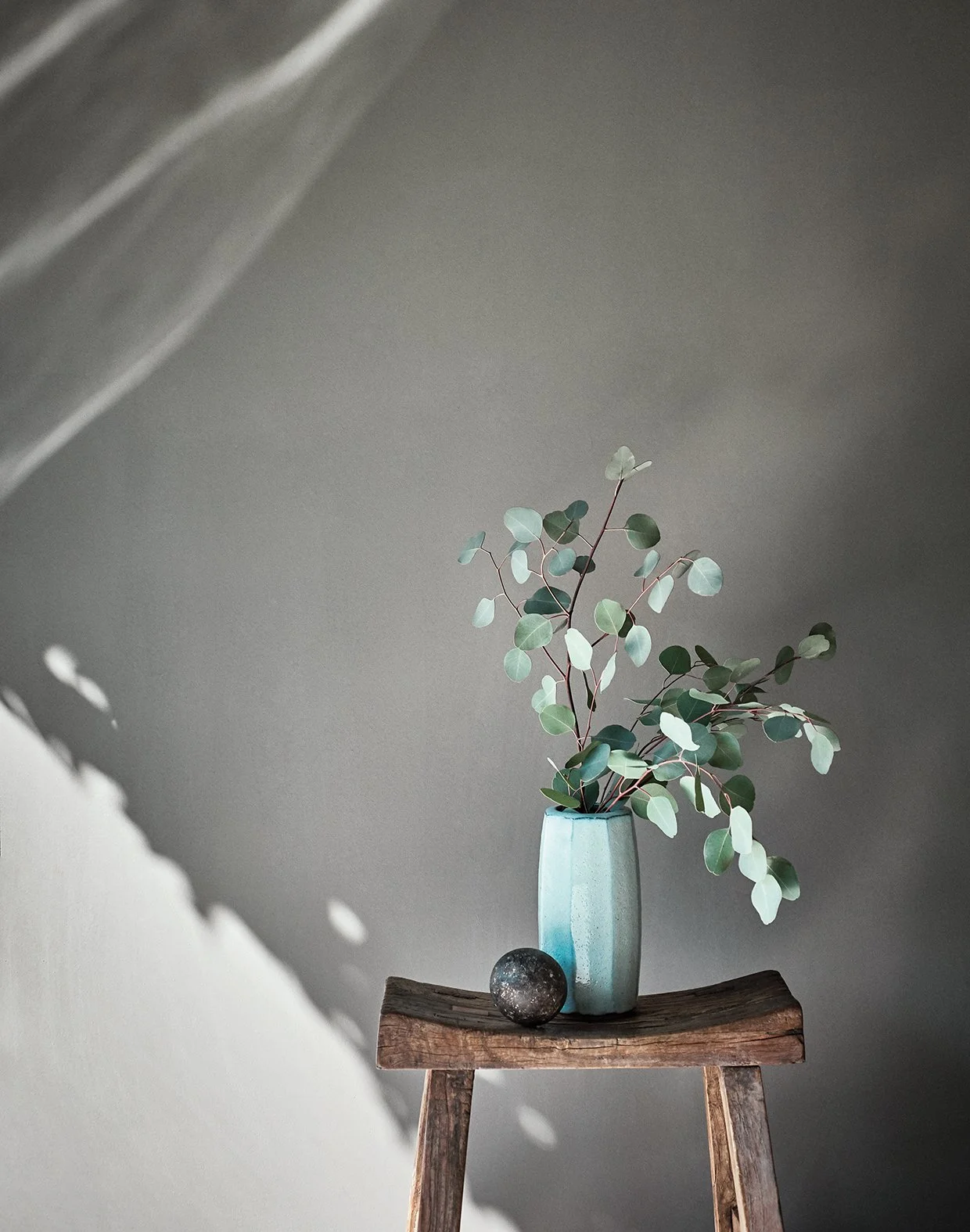 Blumenstrauß mit grünen Blättern in einer blauen Vase auf einem Holzstuhl, Licht wirft Schatten an die Wand.