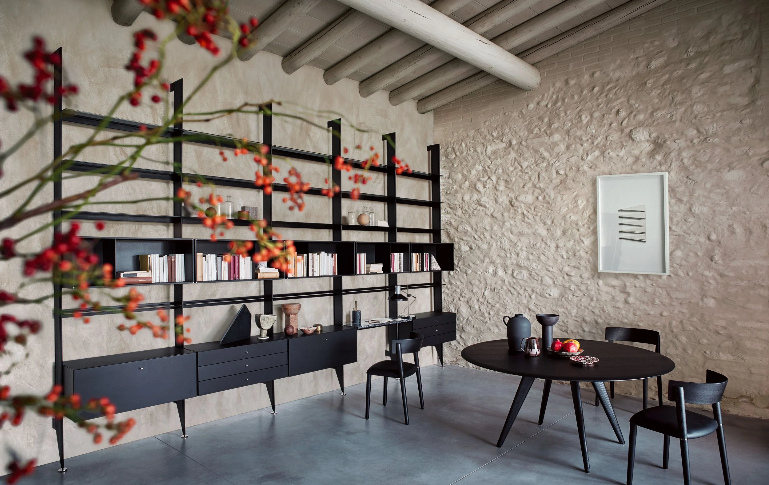 Ein Wohnzimmer mit einer schwarzen Bücherregalwand, einer weißen Steinmauer, Dekorationsobjekten, einem runden schwarzen Tisch mit schwarzen Stühlen und einer Deckenlampe.