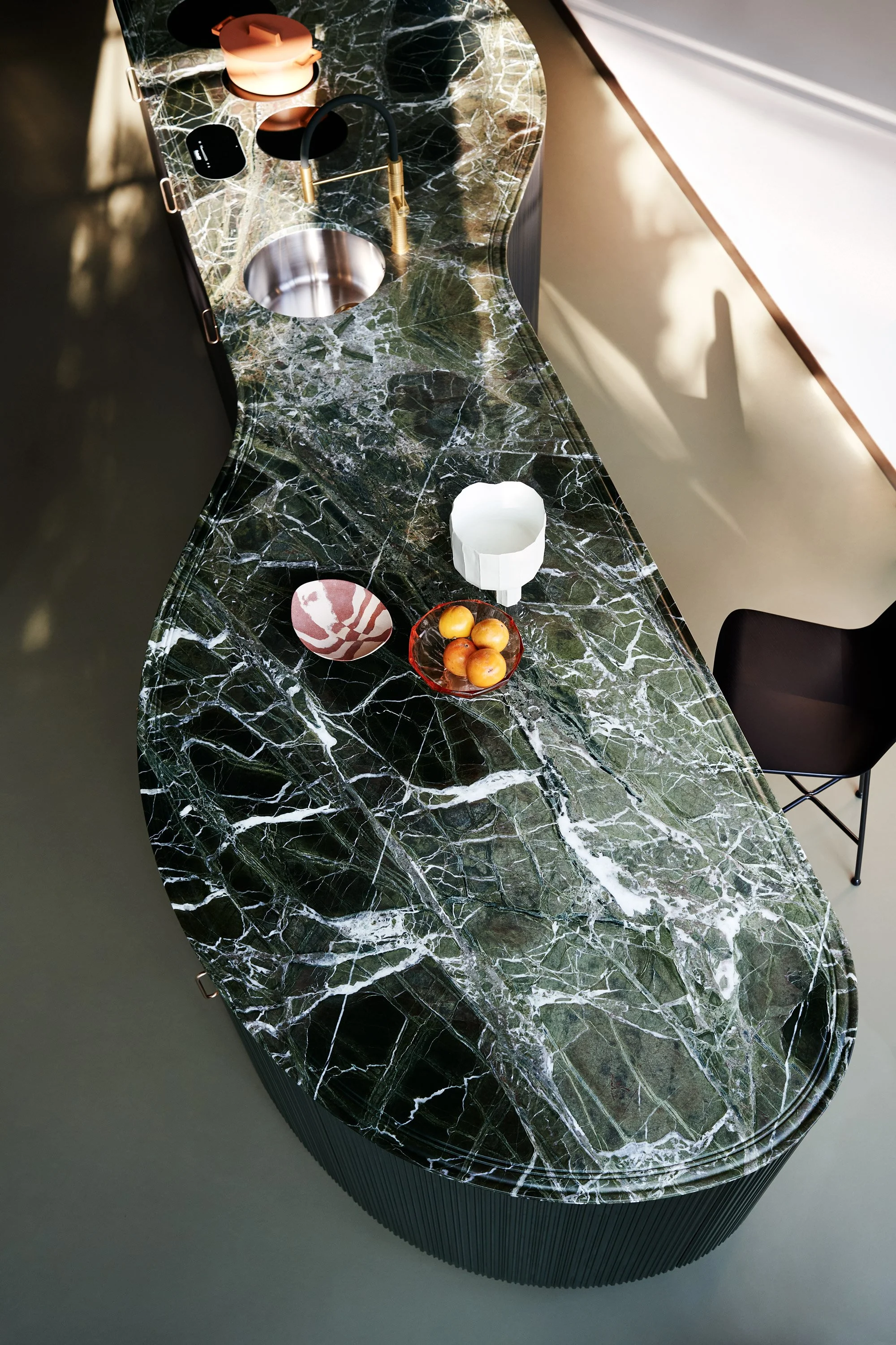Ein Küchenarbeitsplatte aus schwarzem Marmor mit weißen und grünen Adern, auf der sich eine moderne braune Spüle mit goldenem Wasserhahn befindet. Auf der Platte liegen eine Kerze, eine kleine schwarze elektronische Zigarette, eine weiße Vase und ein