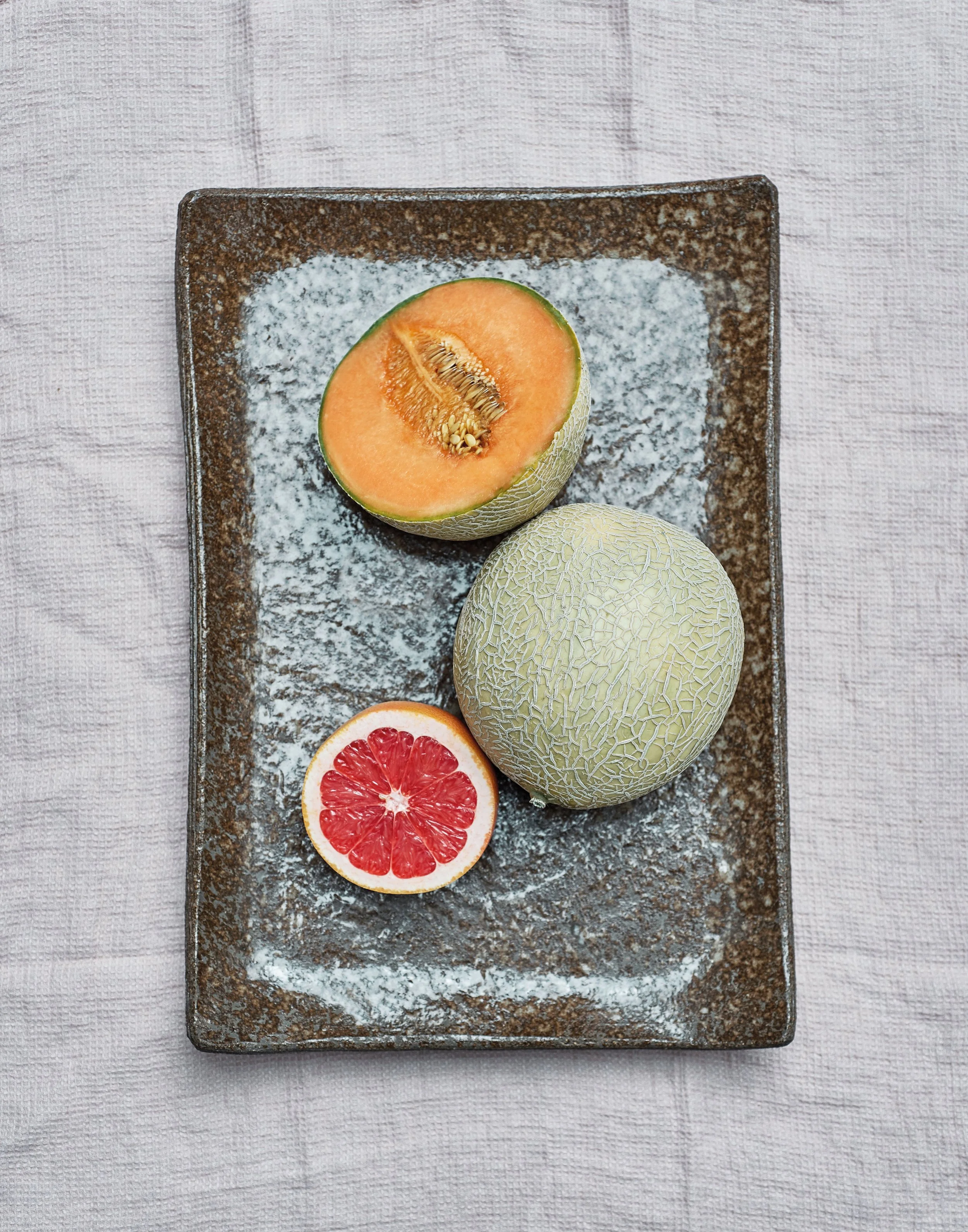 Zwei Melonen, eine halbe Wassermelone und eine halbe Grapefruit auf einem rustikalen braunen Teller, auf einer weißen Stoffdecke.