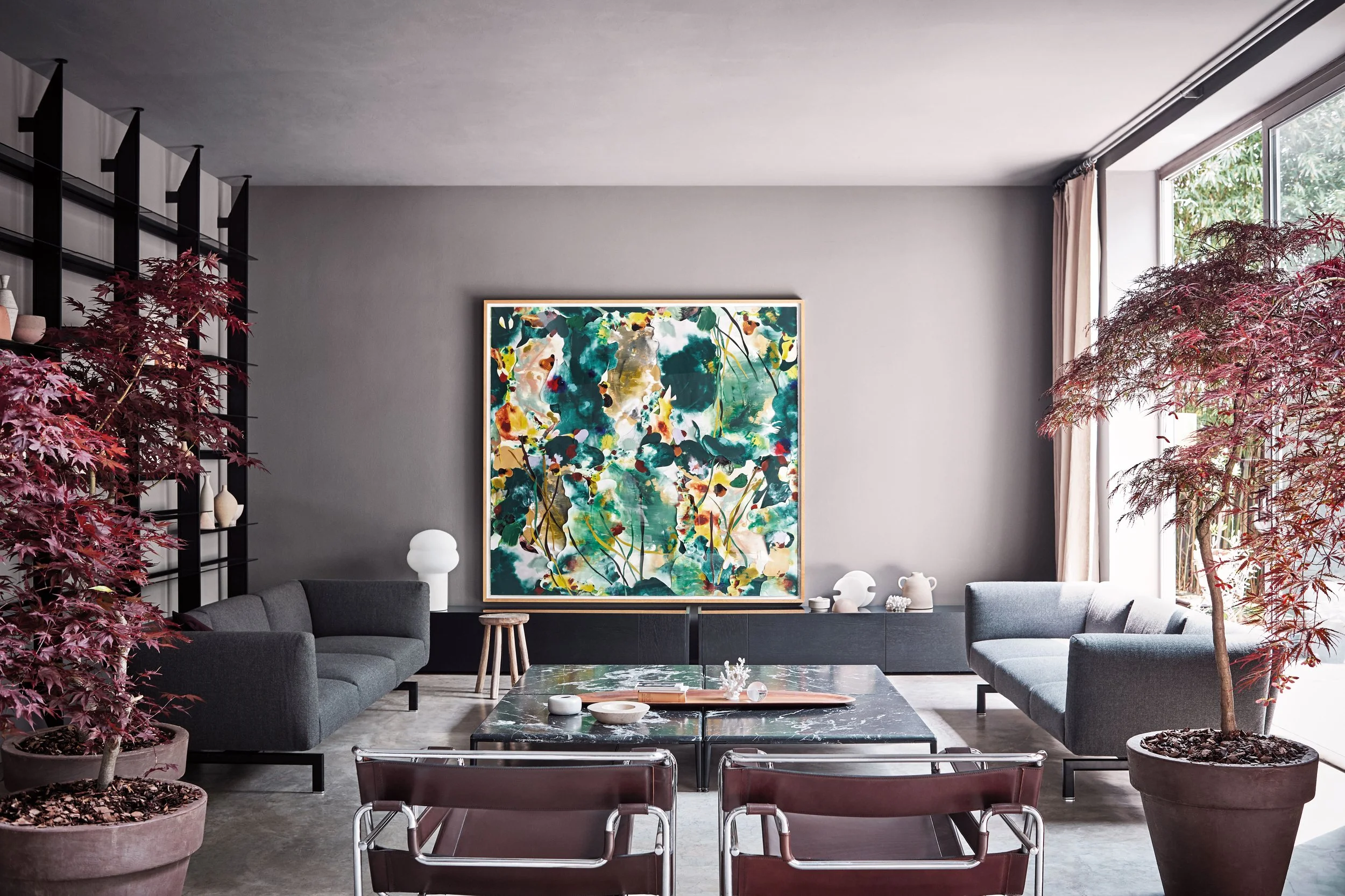 Moderner Wohnzimmerbereich mit grauen sofas, einem abstrakten Kunstwerk an der Wand, großen Fenstern mit Blick nach draußen und roten Topfpflanzen an den Seiten.