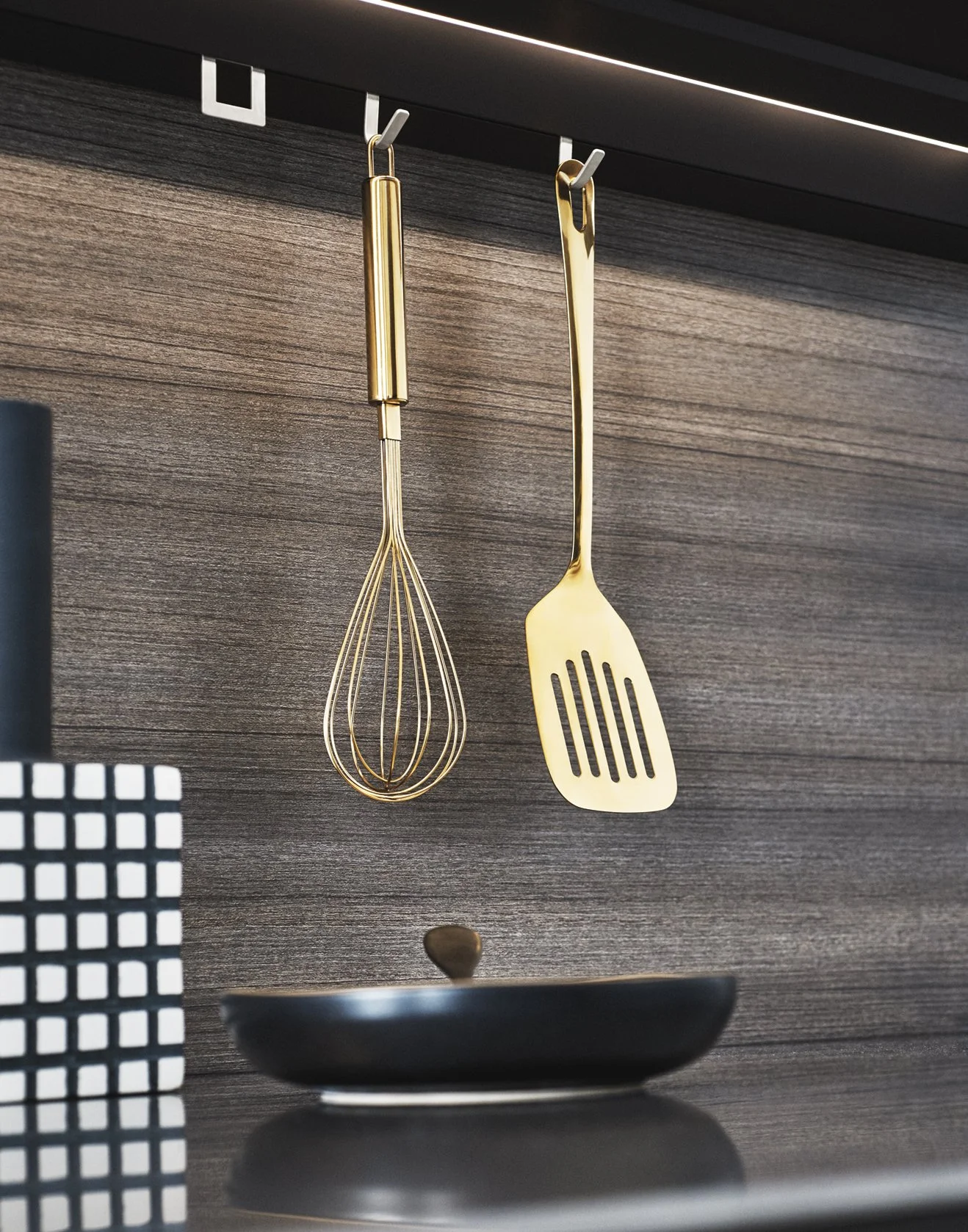 Stilvolle Küchenutensilien hängen an einer eleganten schwarzen Holzwand und schaffen ein modernes Küchendesign mit luxuriösen goldenen Akzenten. Ein goldener Schneebesen, ein goldener Pfannenwender und eine schwarze Pfanne auf dem Herd unterstreichen