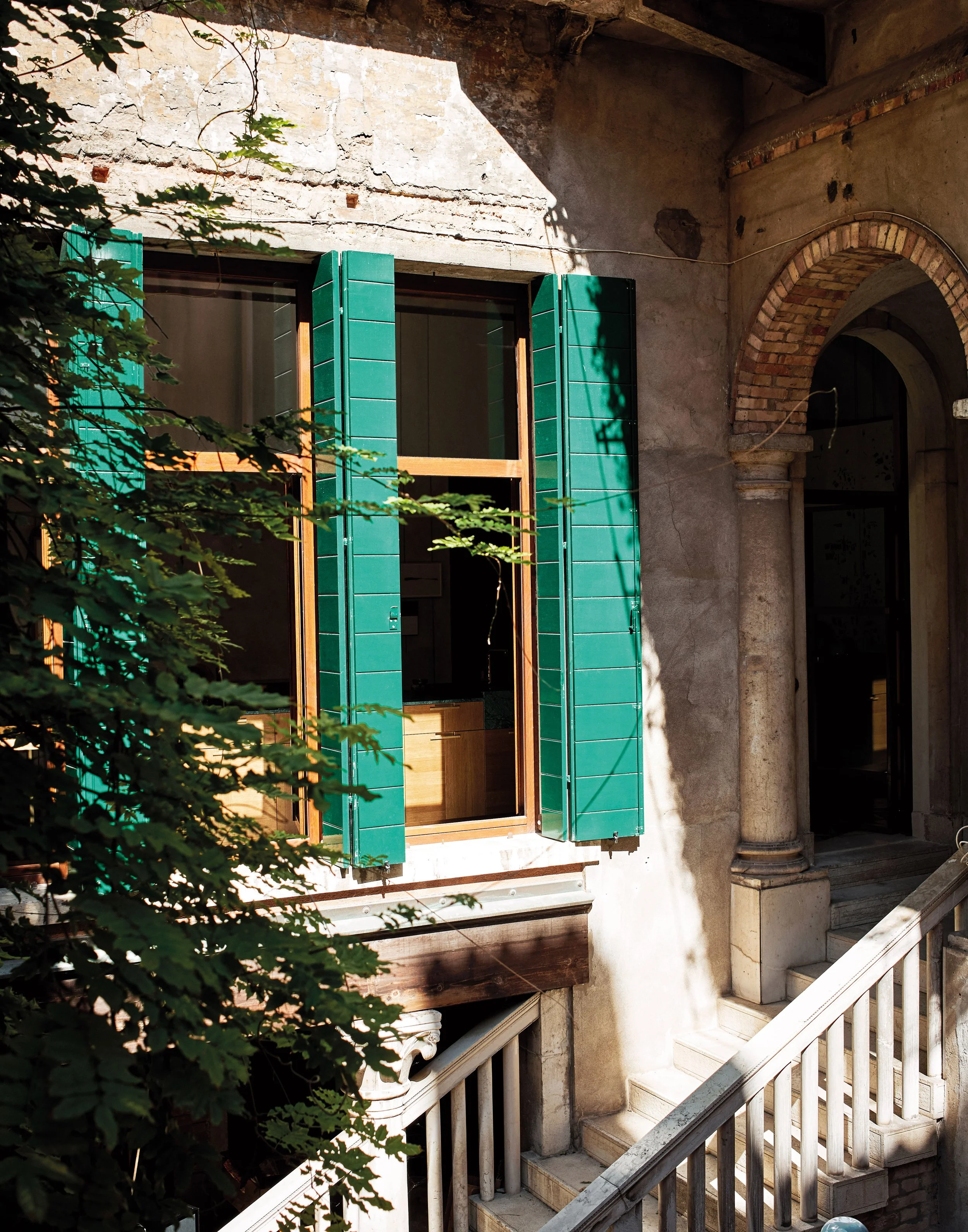 Ein Fenster mit grünen Fensterläden und offener Scheibe, umgeben von einer alten Stein- und Ziegelmauer, mit einem Balkon und einer Treppe mit Lambert-Stil-Design.