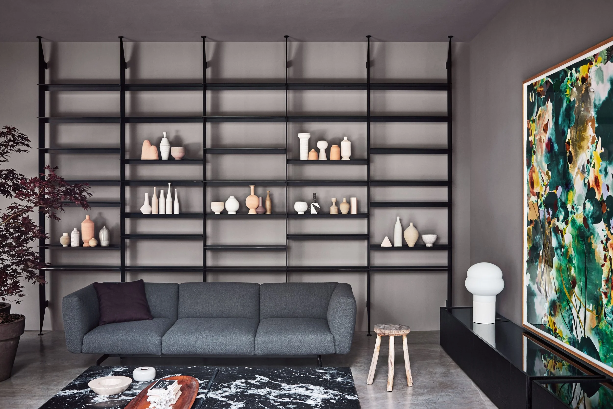 Ein modernes Wohnzimmer mit grauem Sofa, schwarzen Bücherregalen an der Wand, bunten Vasen und einer großen bunten Kunstoptik an der Wand.