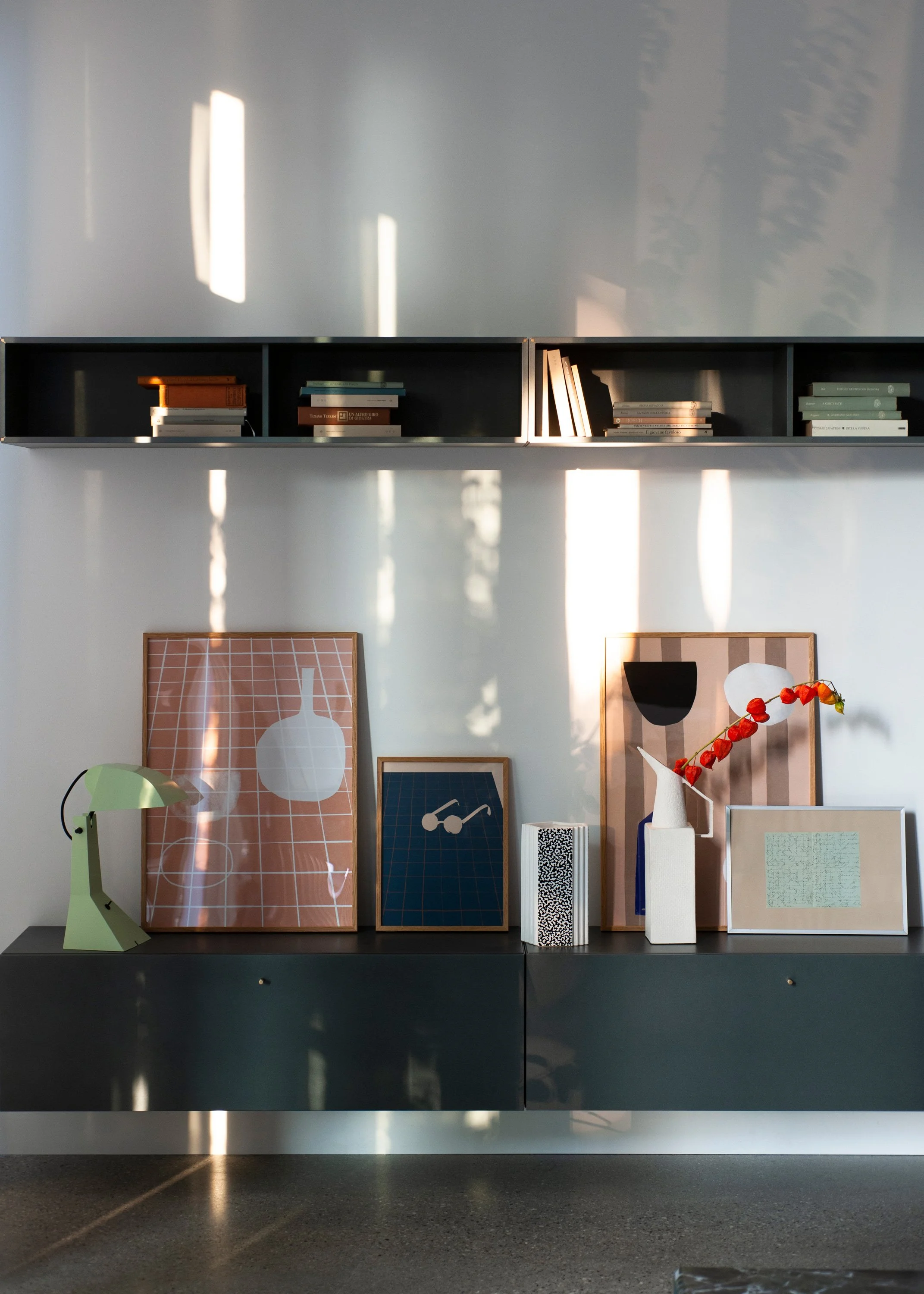 Wandregal mit Büchern, Dekorationen und Kunstwerken in einem Raum mit Sonnenlicht, das durch Fenster fällt.