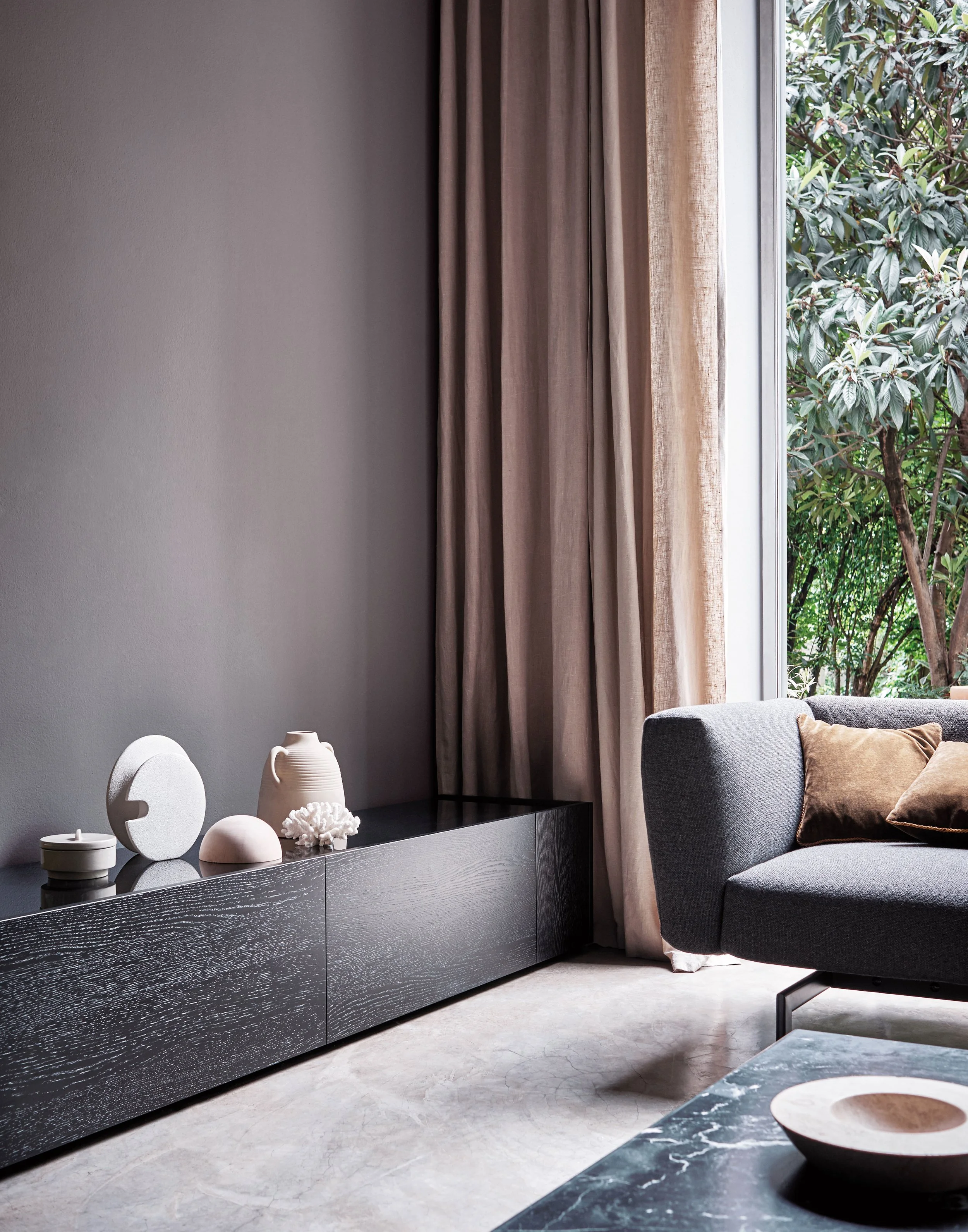 Ein modernes Wohnzimmer mit dunklem Sideboard, weißen Dekorationsgegenständen, grauem Sessel, großen Fenstern mit braunen Vorhängen und natürlichem Licht, Blick auf grüne Bäume draußen.
