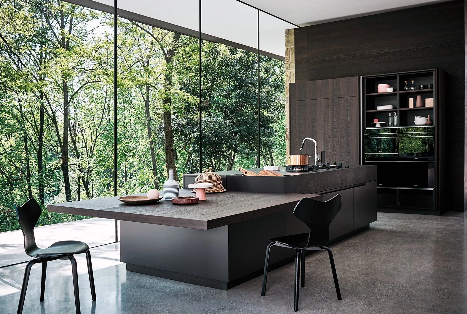 Moderne Küche mit großen Fenstern, Blick auf einen grünen Wald, dunkle Einrichtung, Esstisch mit schwarzen Stühlen und Küchenarbeitsfläche mit Geschirr und Küchenutensilien.