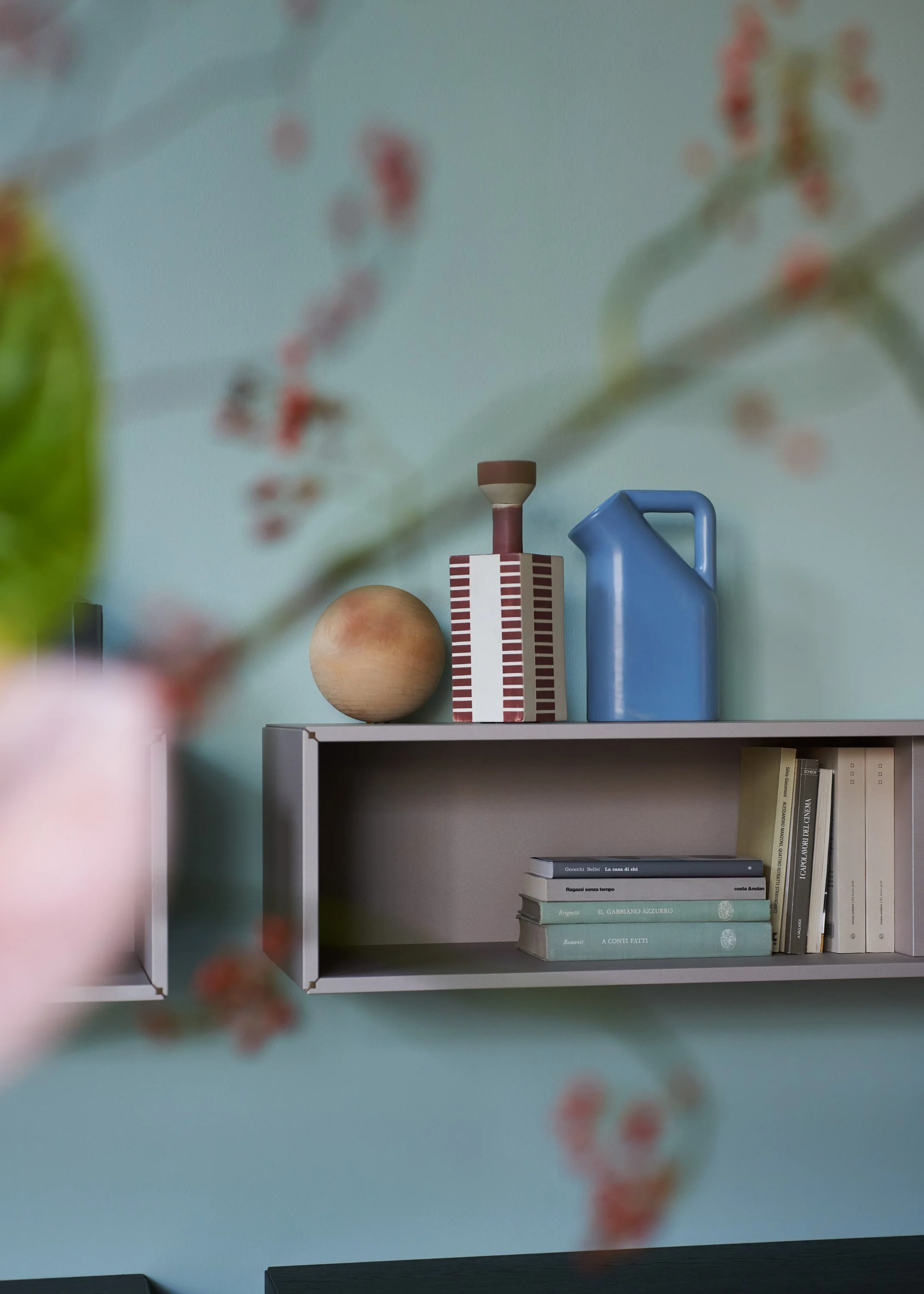Blick auf eine weiße Bücherregal mit buntem Geschirr, Büchern und Dekorationsgegenständen vor einer blauen Wand, der Blick ist durch Blumen im Vordergrund etwas unscharf