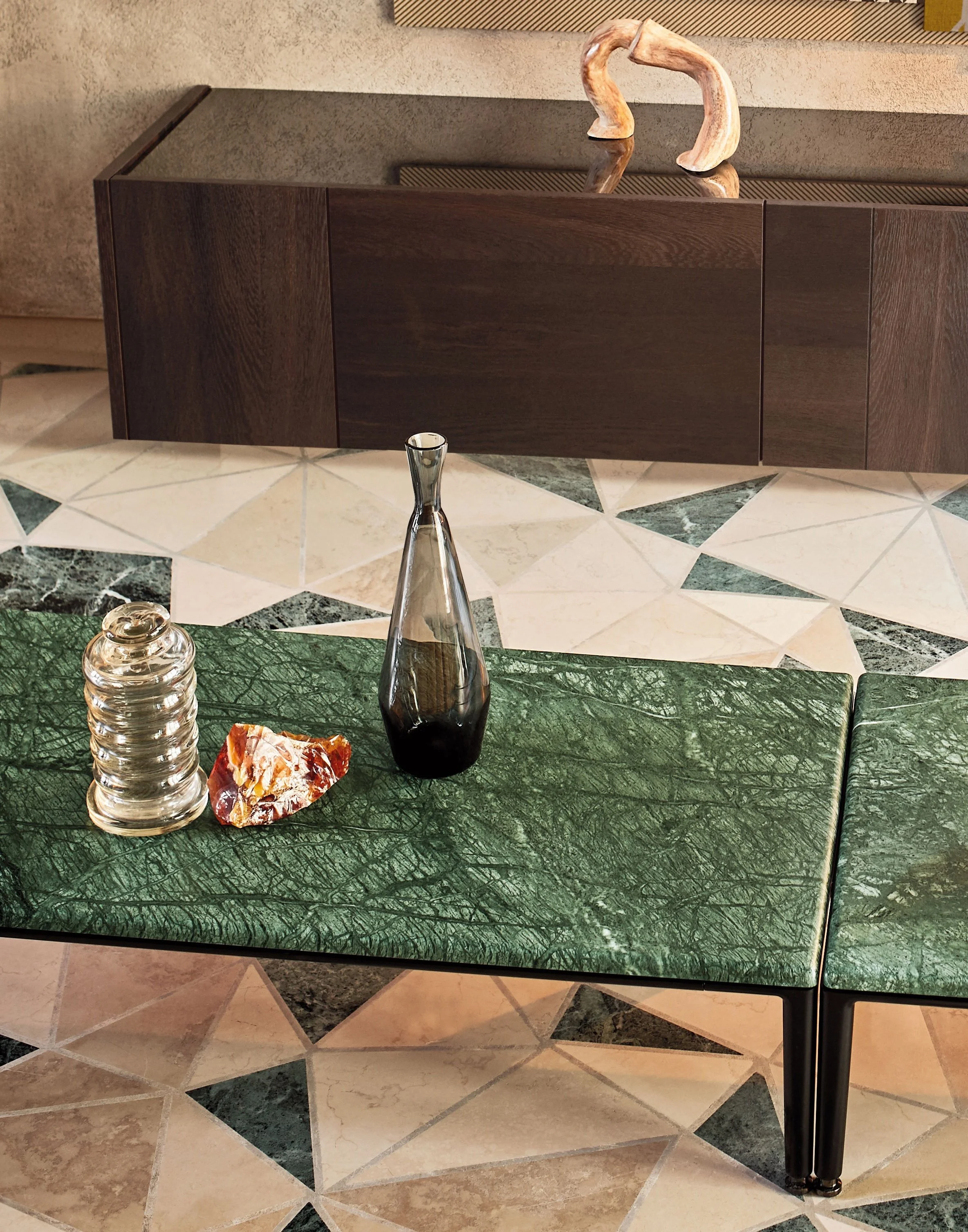 Ein dunkelgrüner Marmor-Tisch mit einer transparenten Glasvase, einem kleinen Glasbehälter und einer farbigen Muschel. Im Hintergrund eine dunkelbraune Holzwand mit Dekoration und ein Schrank aus dunklem Holz über einem hellen und dunklen Fliesenbode