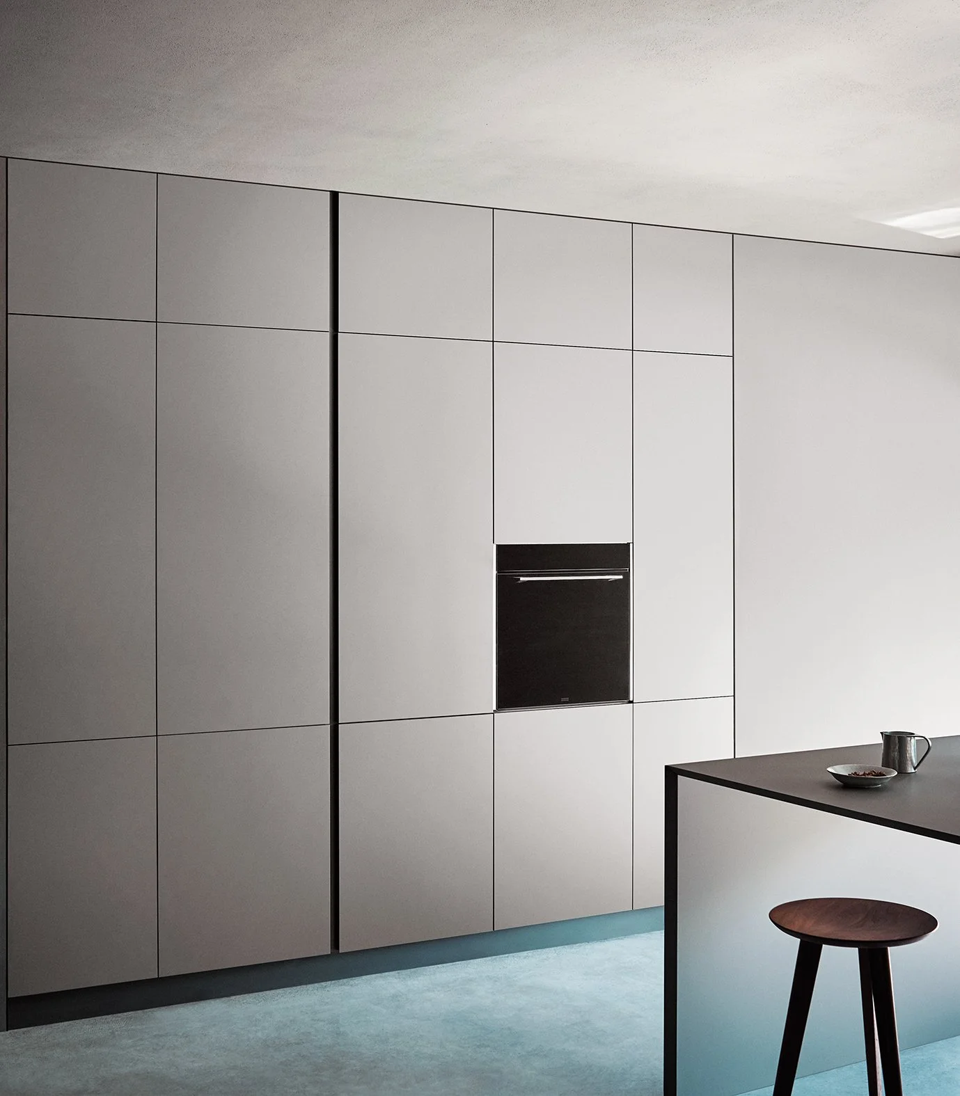 Moderne, minimalistische Küche mit weißen Schränken, eingebautem schwarzen Backofen, dunklem Tisch und Hocker, grauem Boden, weißer Wand und Decke.