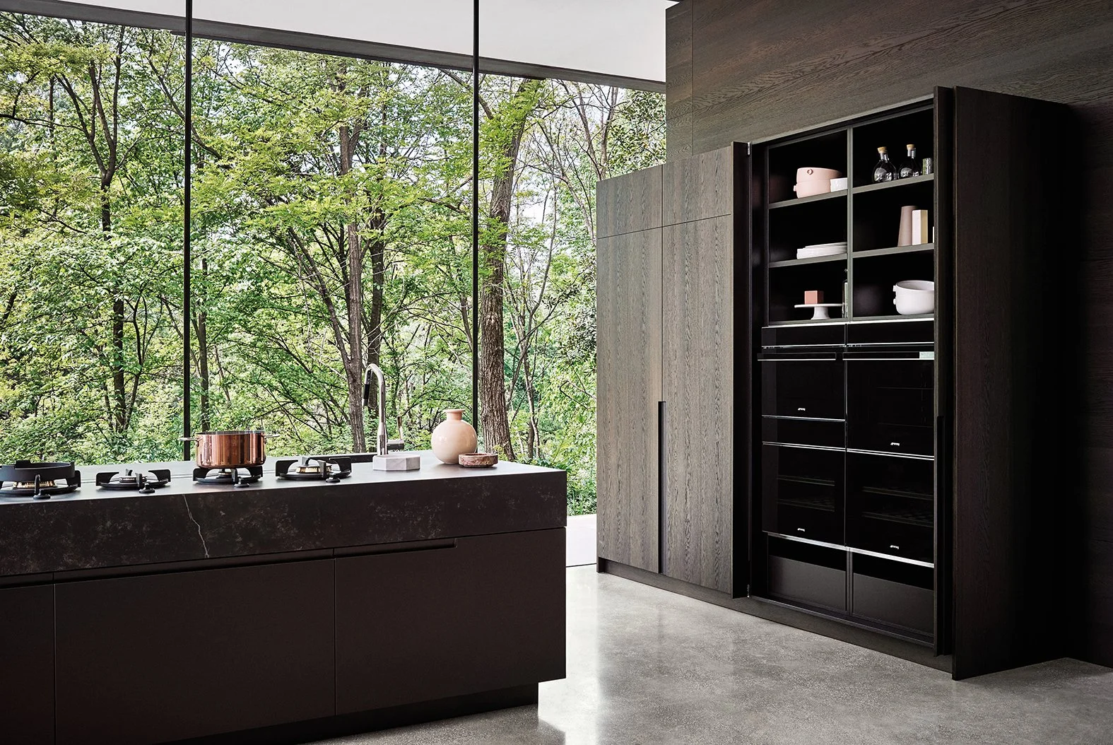 Moderne Luxusküche mit eleganter Kücheninsel und integriertem Kochfeld vor großen Fenstern mit Blick auf grüne Bäume. Die stilvollen schwarzen Wandregale, hochwertigen Küchenmöbel und die natürliche Beleuchtung schaffen ein modernes und minimalistisc