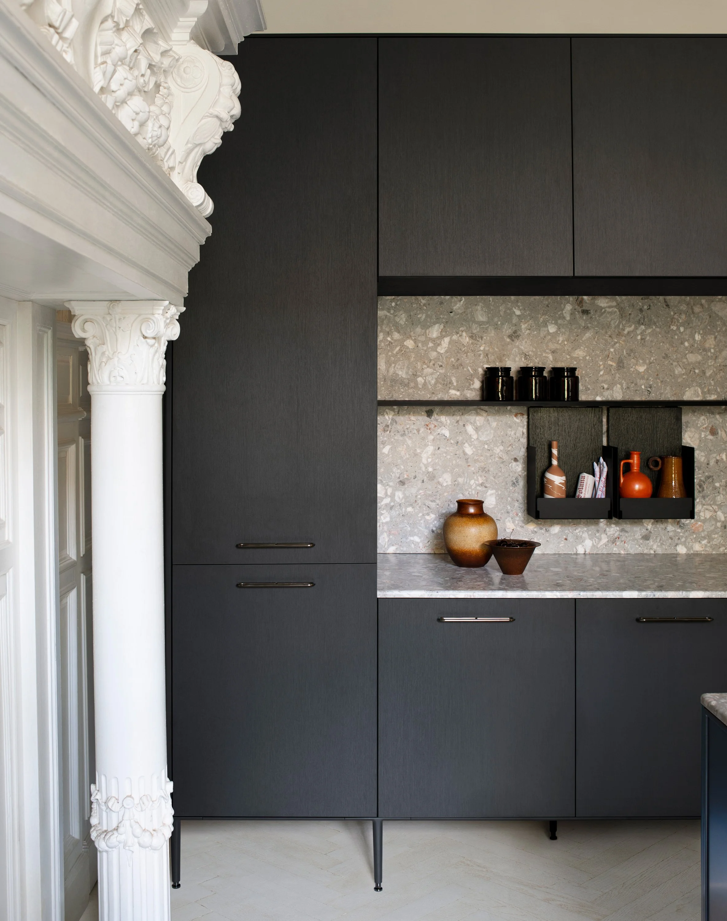 Moderne schwarze Küchenschränke mit Mittelputz aus Terrazzo, dekorative schwarze Behälter auf offener Wandregal, antike weiße Säule mit kunstvoller Verzierung im Vordergrund.