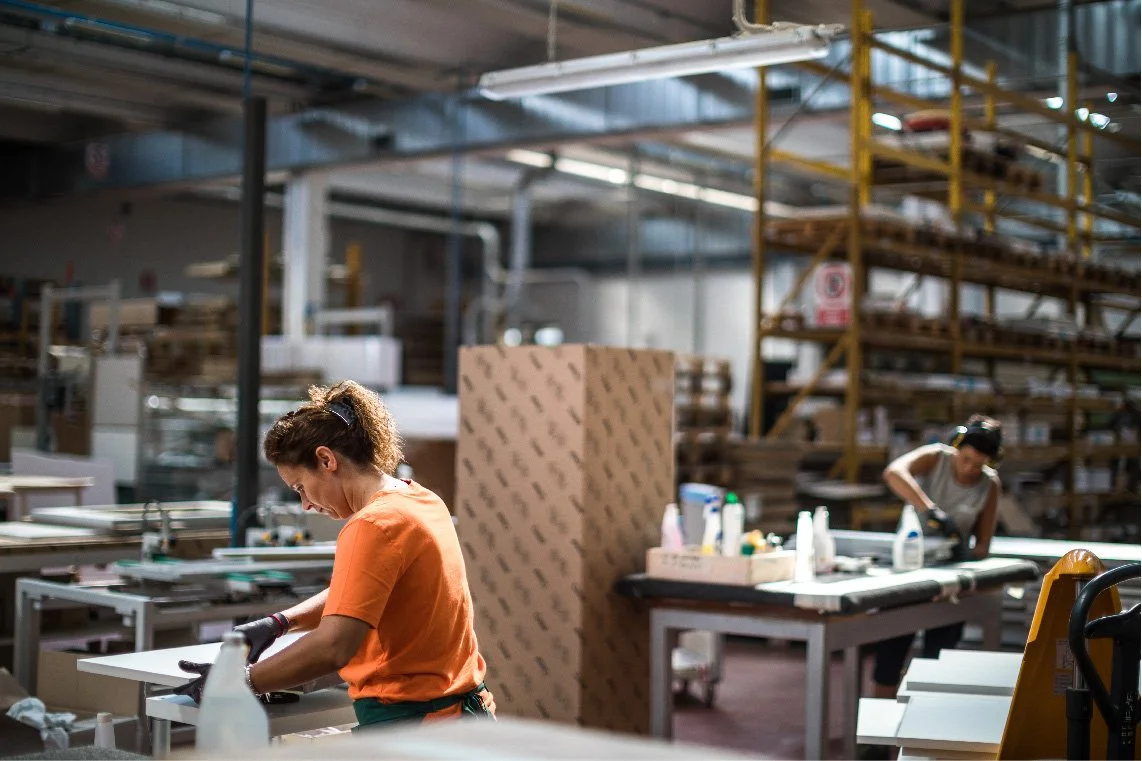 Zwei Frauen arbeiten in einer Lagerhalle oder Fabrik, die mit Regalen voller Materialien gefüllt ist.