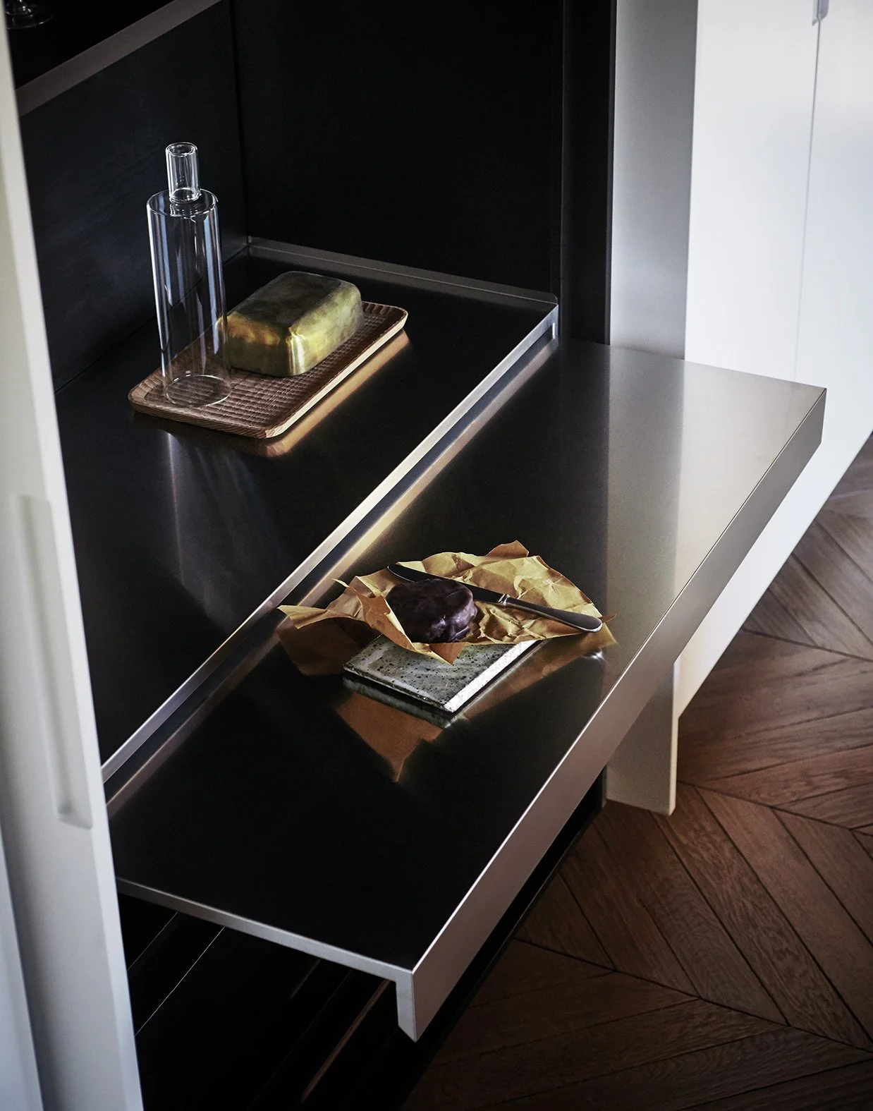 Ein moderner Küchenbereich mit schwarzen und weißen Oberflächen. Auf einem schwarzen Metalltisch liegen eine Schoko-Donut auf Papier, ein Messer und ein Schneidebrett. Im Hintergrund steht eine Glasflasche und eine Seife auf einem kleinen Tablett.
