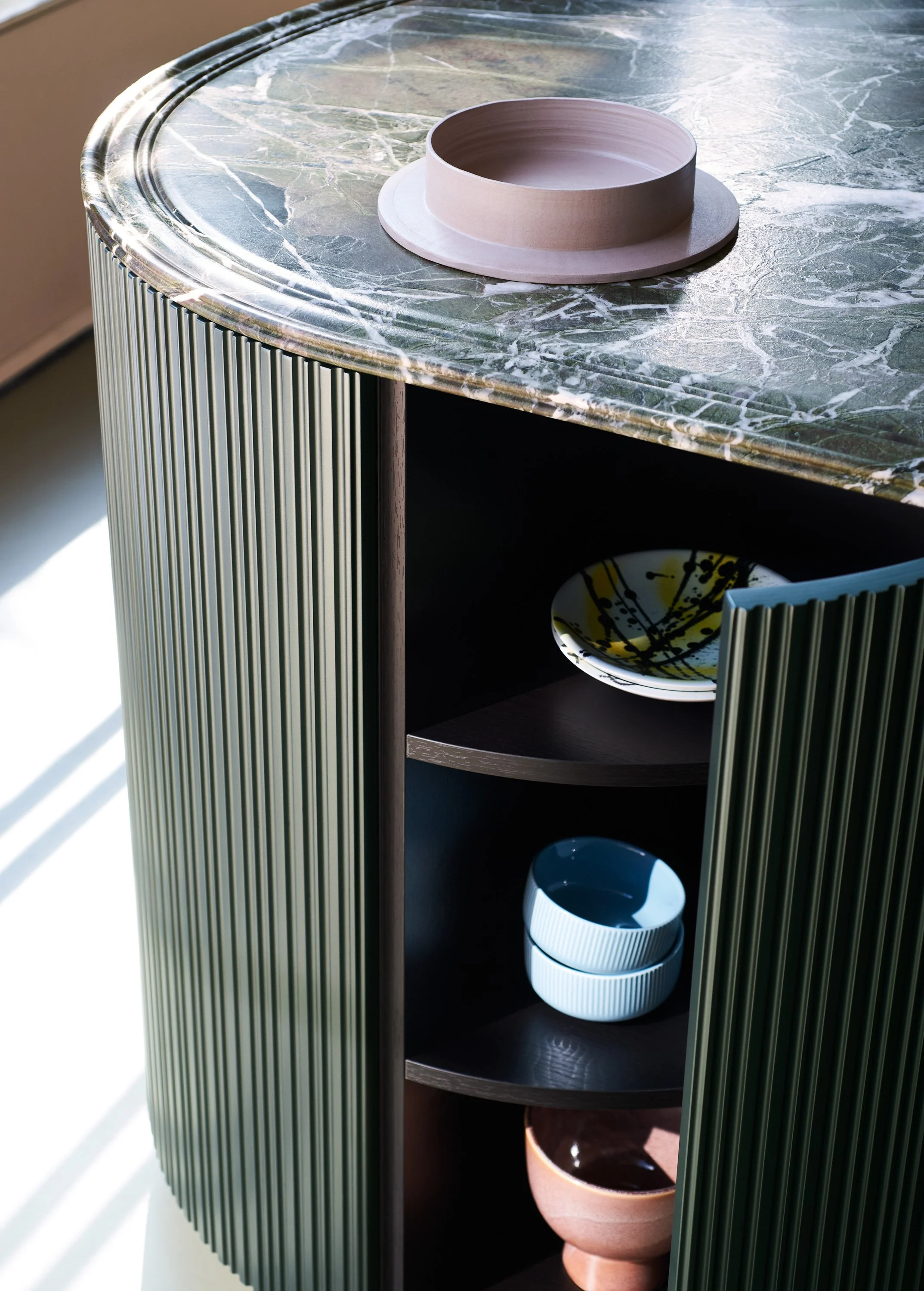 Ein Tisch mit grünem Marmor-Design, darauf ein runder, schlichter Kerzenschirm und darunter ein Schwarzregal mit mehreren Schalen, darunter eine weißen mit schwarzen Linien und eine terrakottafarbene.